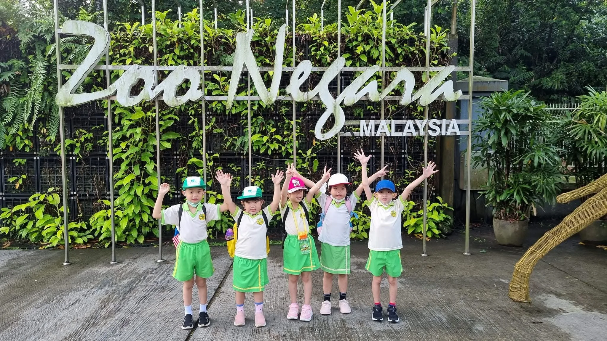 Five children in green shorts and hats with backpacks posing in front of Zoo Negara Malaysia entrance sign surrounded by greenery.