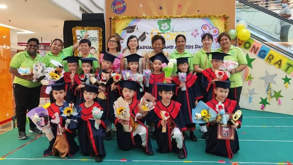 Group of children in graduation gowns holding flowers with adults on a stage with Congrats banner.