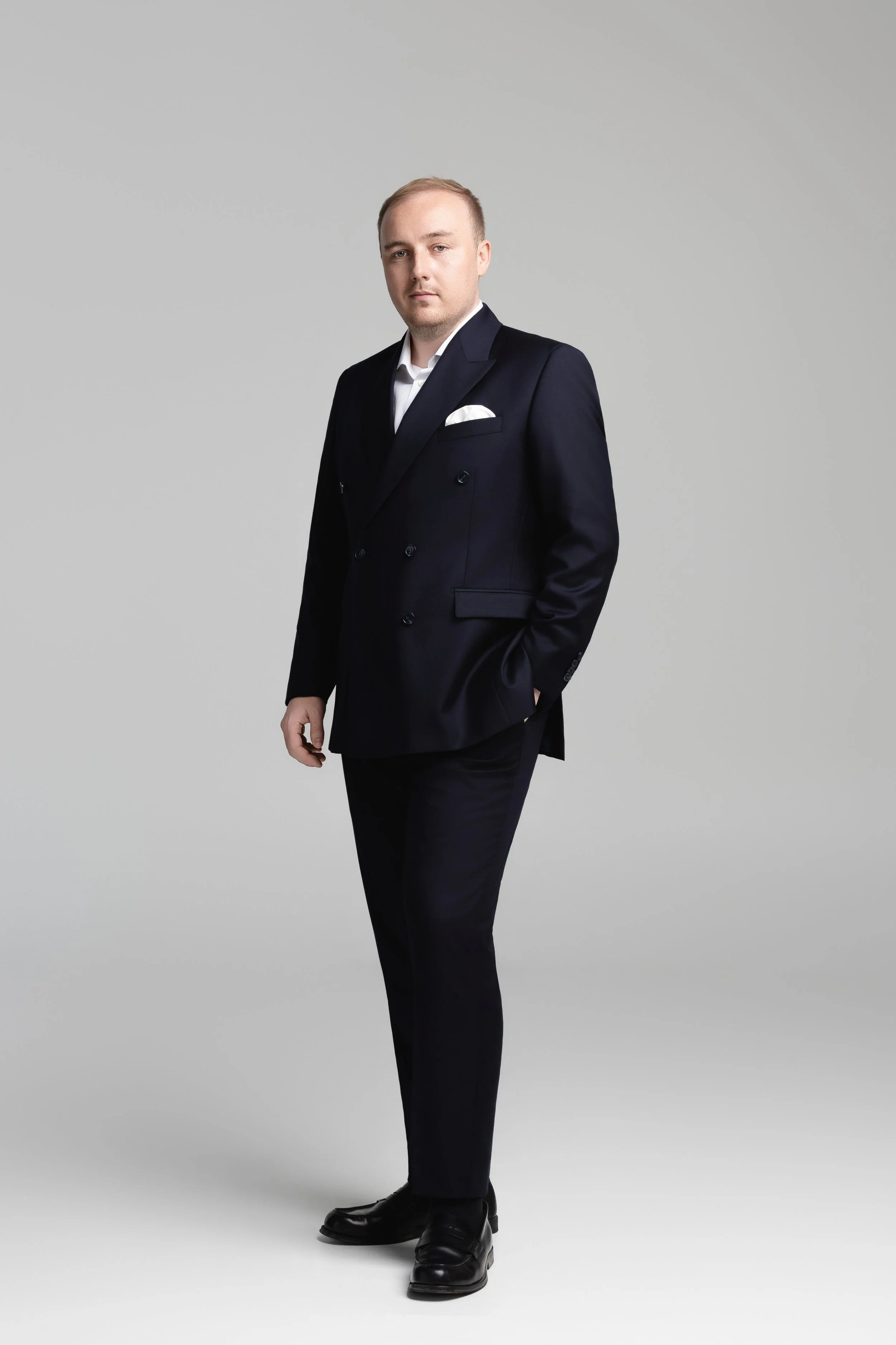 A young man in a dark suit, white shirt, and black shoes standing against a plain gray background, with a serious expression and his hands at his sides.