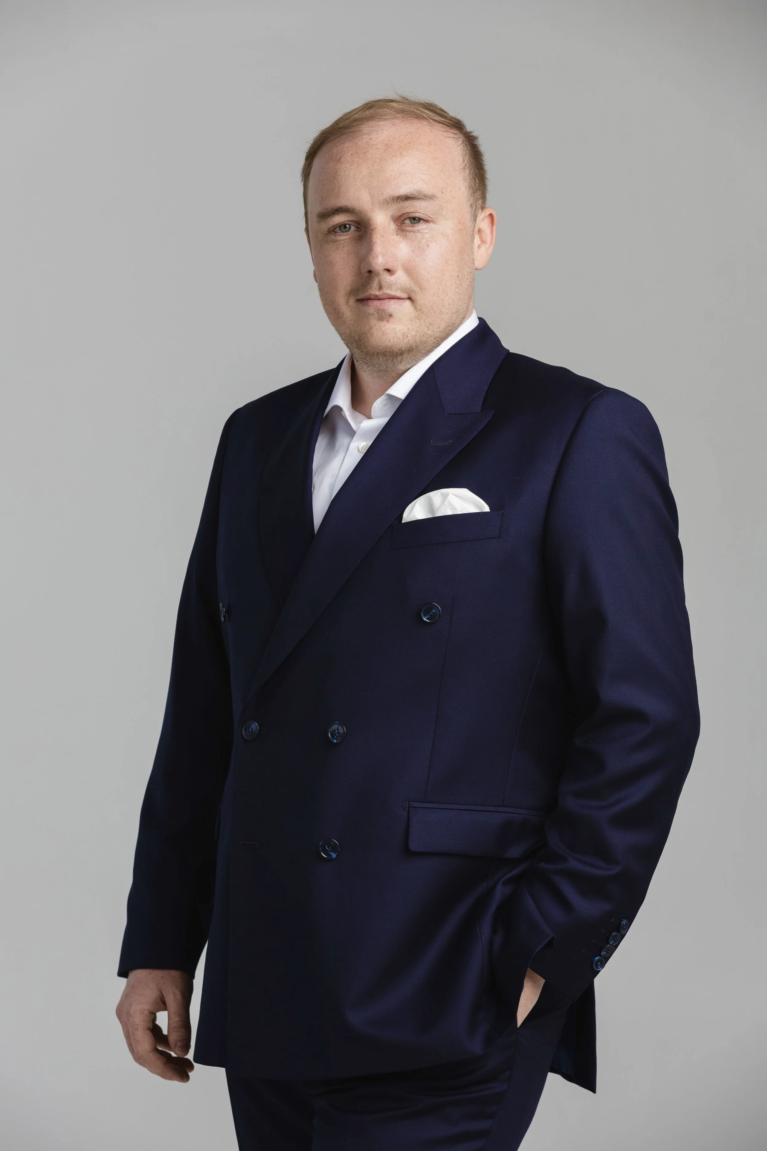 A young man wearing a navy blue double-breasted suit with a white shirt and pocket square, standing against a plain gray background.