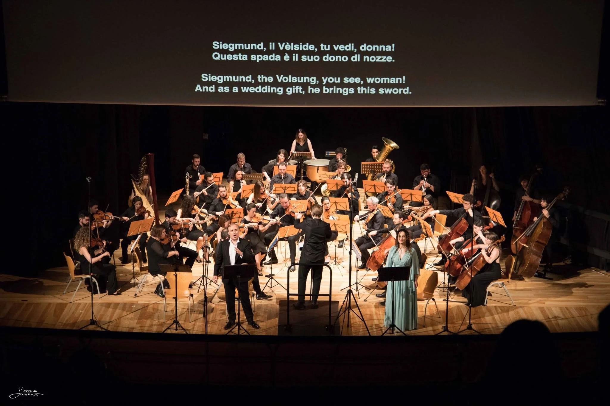 Symphony orchestra performing on stage with a conductor in front. A screen above displays lyrics in Italian and English about Siegmund, the Volsung, and a wedding sword.