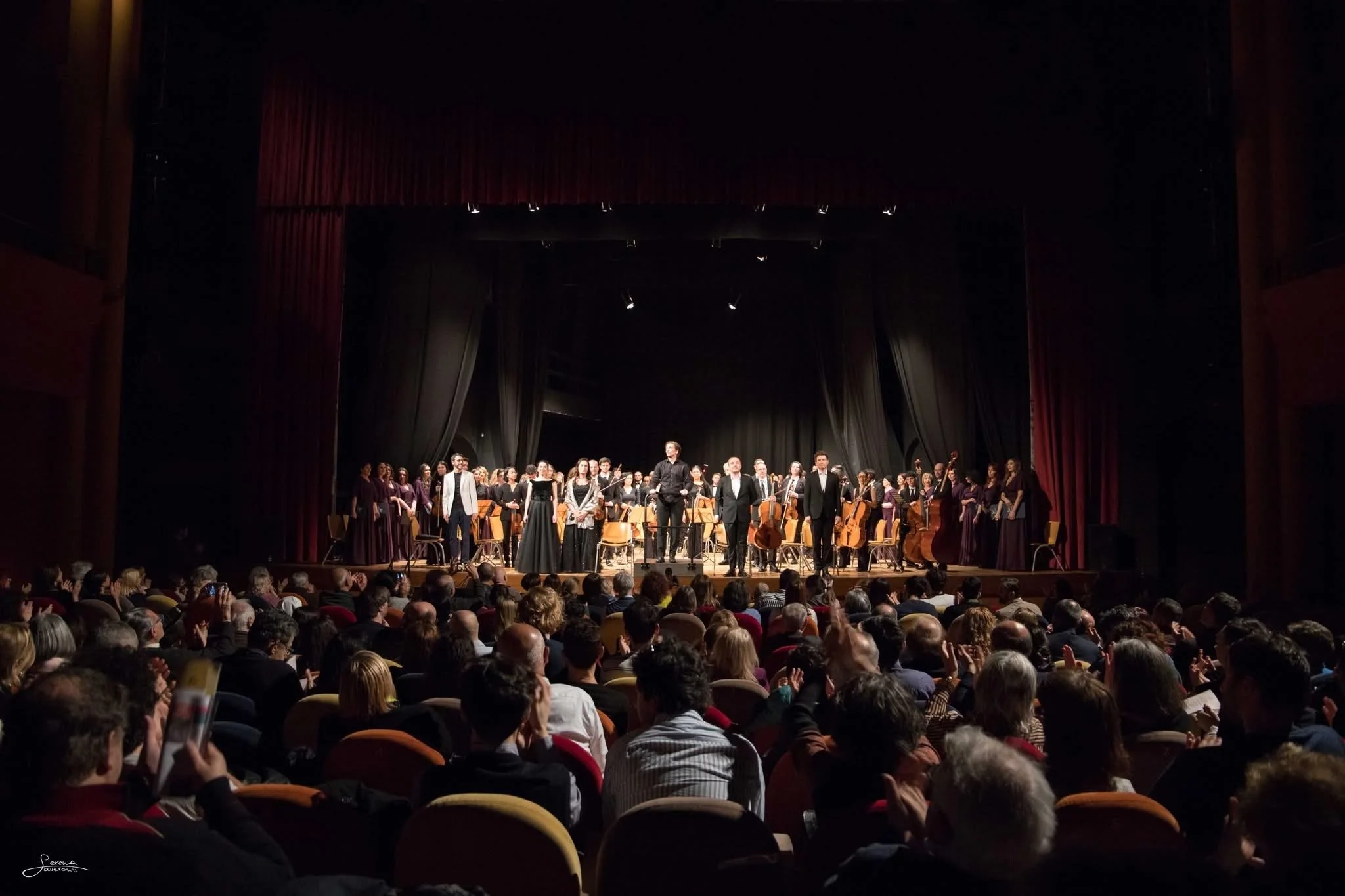 An orchestra performing on a stage in a theater with an audience watching. The stage has musicians with string instruments, and a conductor standing in front. The audience is seated in rows with some people clapping and taking photos.