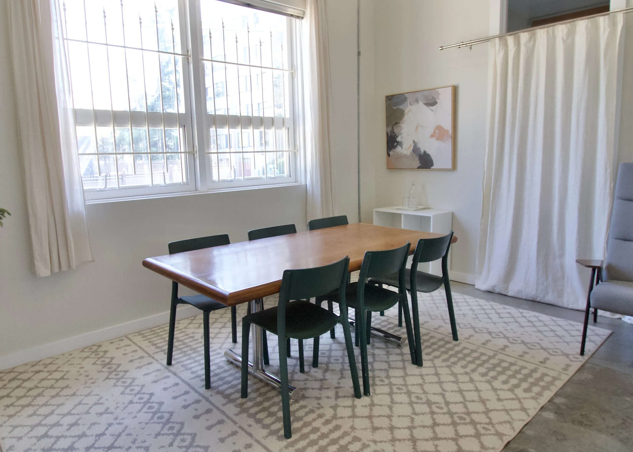 A dining area with a wooden table surrounded by six black chairs, positioned on a patterned rug. Behind the table is a large window with white curtains, allowing natural light to illuminate the room. On the wall near the window, there is a modern abstract painting and a small white side table with a water bottle and glass.