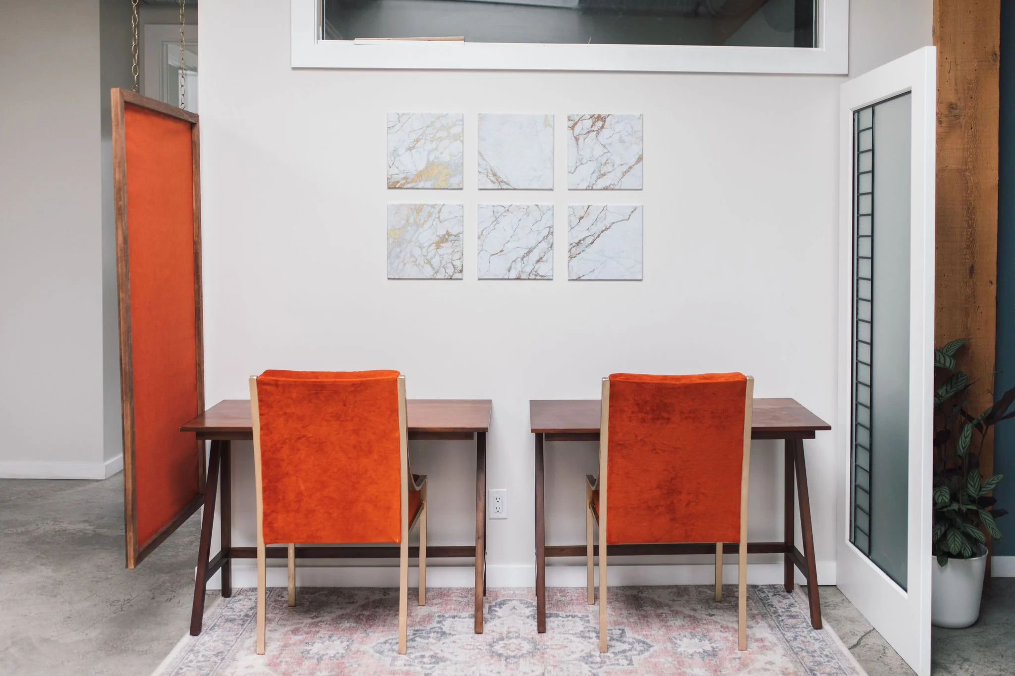 A modern dining area with a wooden table and six orange upholstered chairs. The wall behind features six pieces of marble-patterned artwork arranged in two rows. To the left, there is a wooden partition with a fabric insert, and to the right, part of a door with a glass panel and black grid design. The floor is covered with a patterned rug under the table.