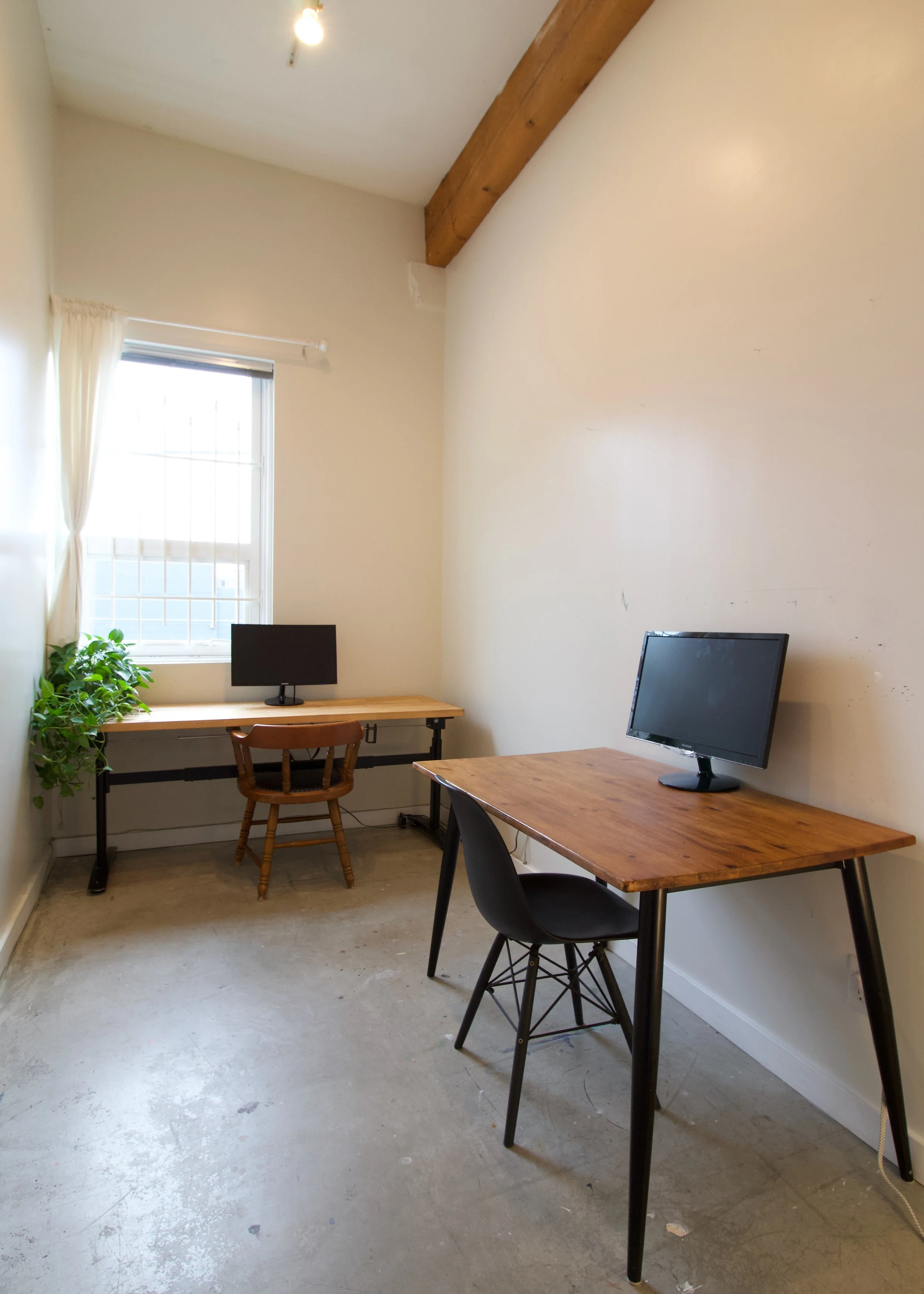 A room with two wooden desks, each with a computer monitor, a chair, a window with curtains, a brick wall, and a wooden beam on the ceiling.