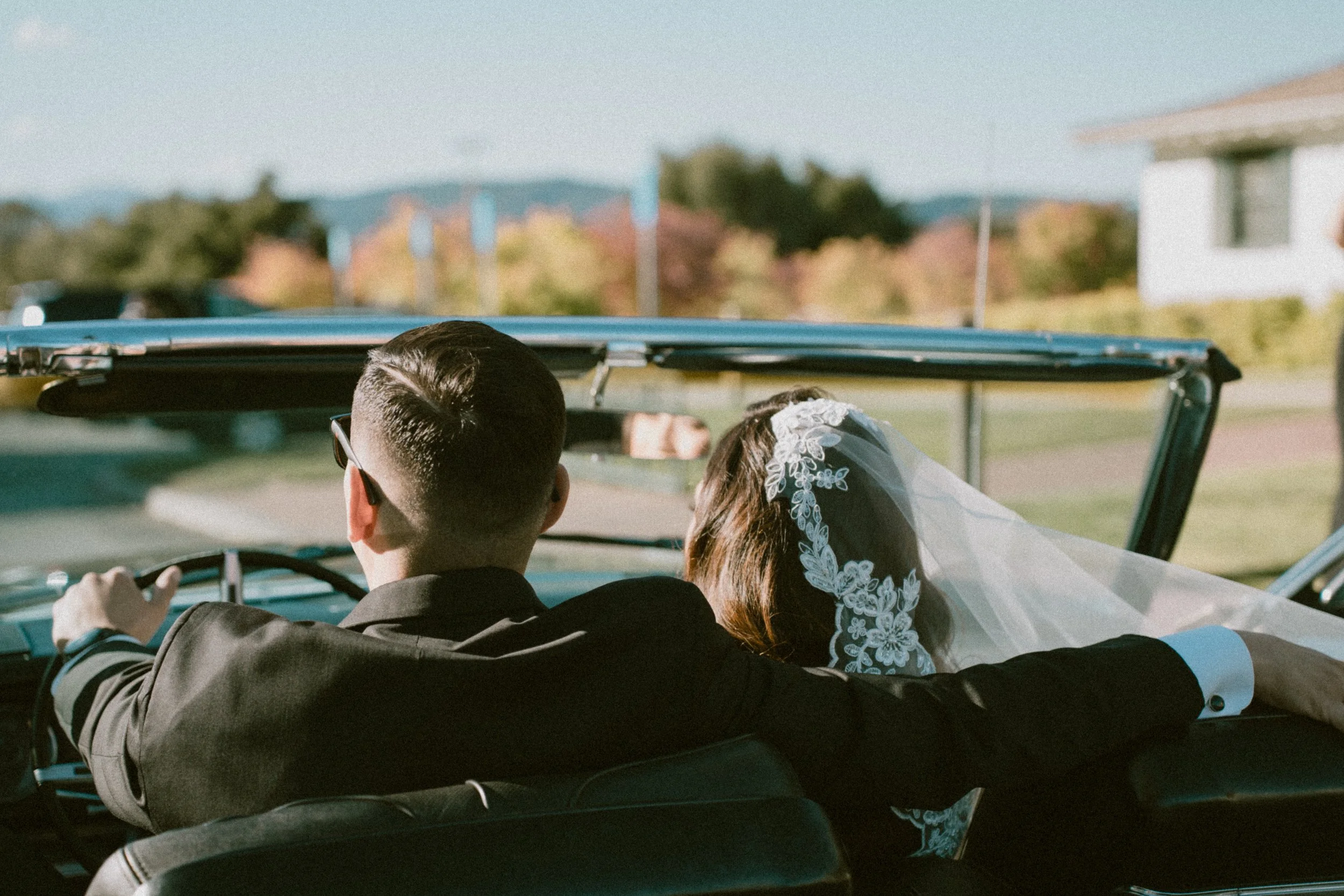 A newlywed couple sitting in the front seat of a vintage convertible car, with the groom driving and the bride wearing a lace wedding veil, enjoying a scenic drive on a sunny day.
