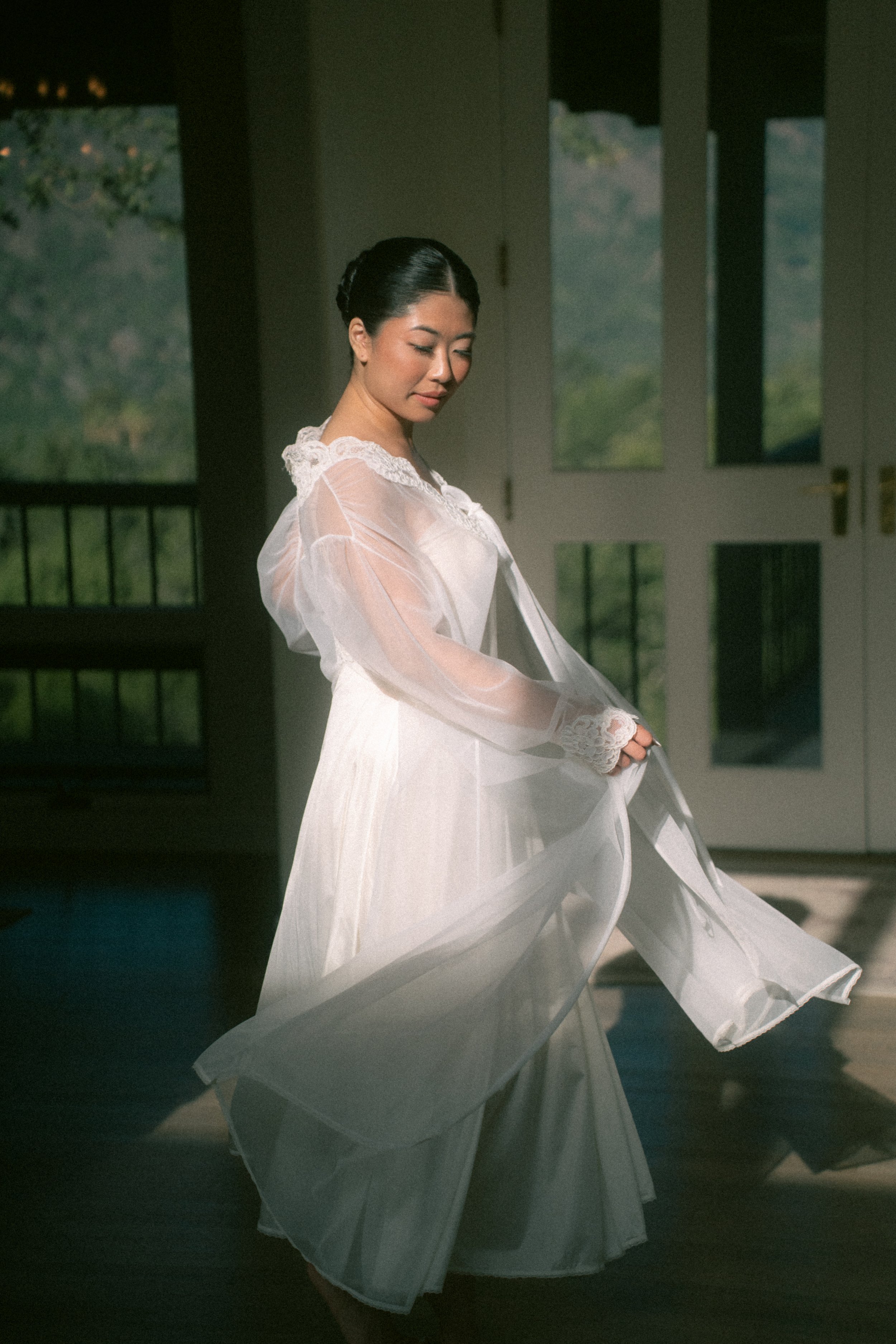 A woman in a flowing white dress, standing indoors near a door with glass panels, with sunlight casting soft shadows.