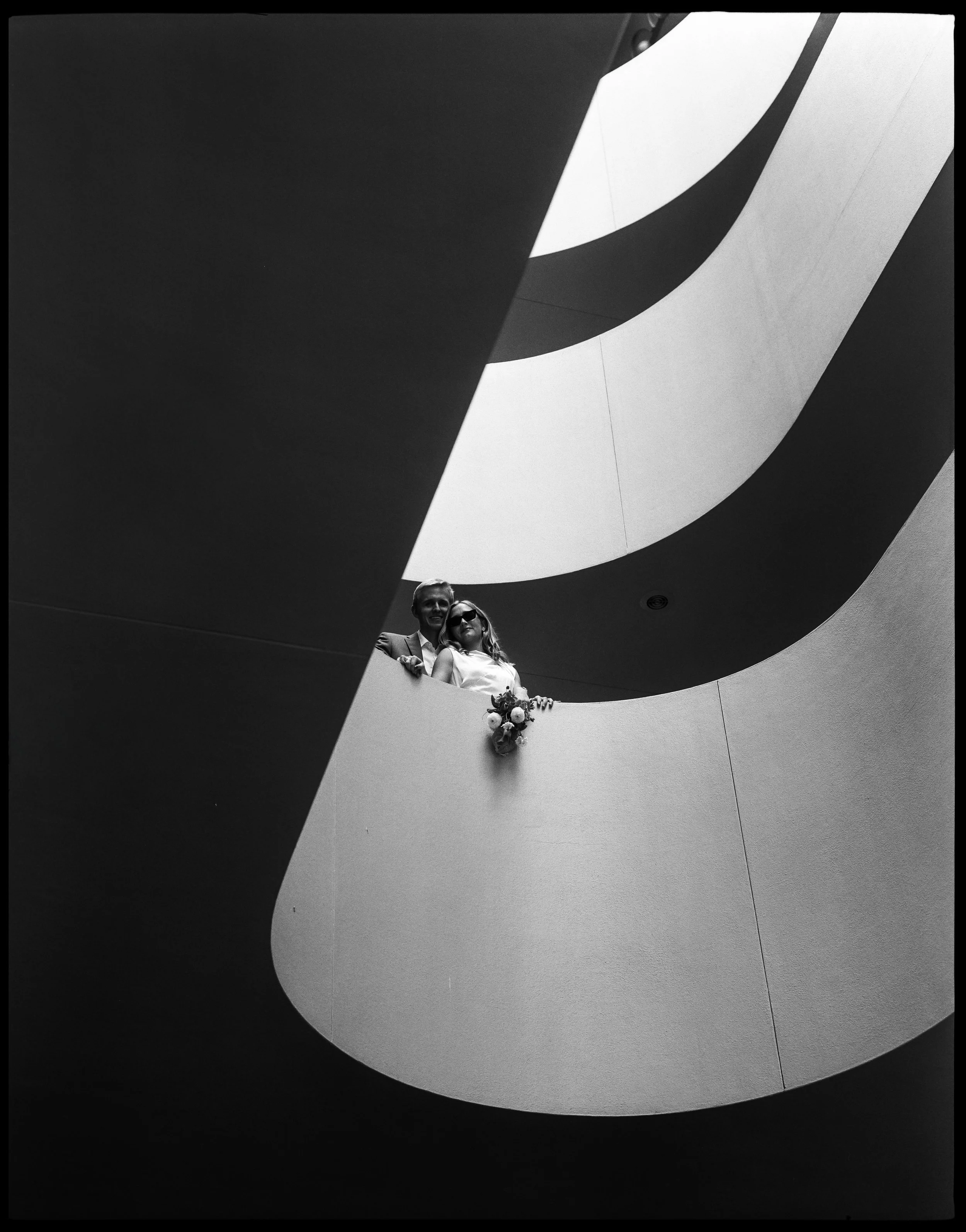 A black and white photo of a couple standing on a modern curved balcony, looking down from a spiral staircase or atrium.