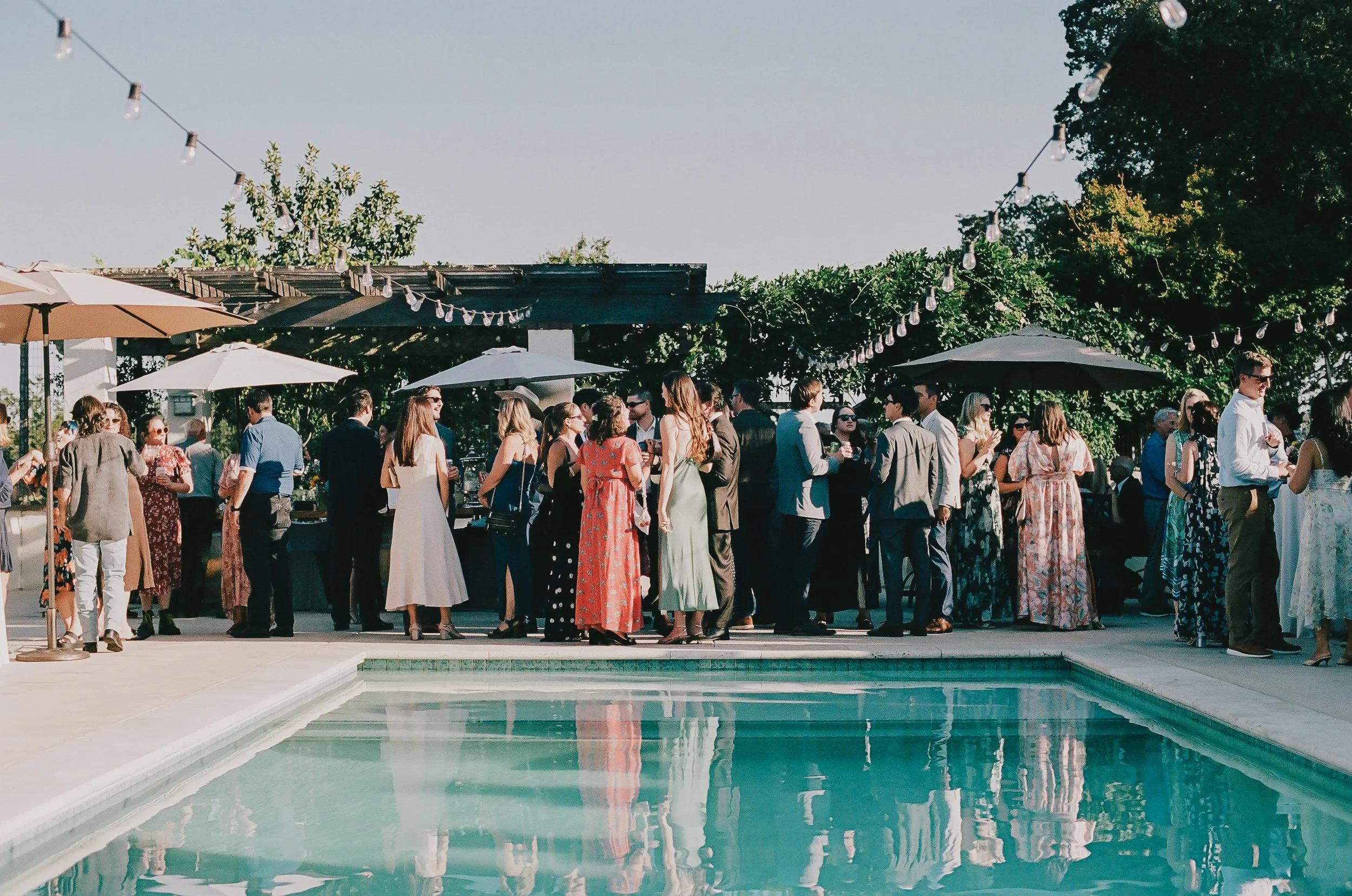 A group of people in formal attire at an outdoor poolside gathering during daytime, with string lights, umbrellas, and trees in the background.