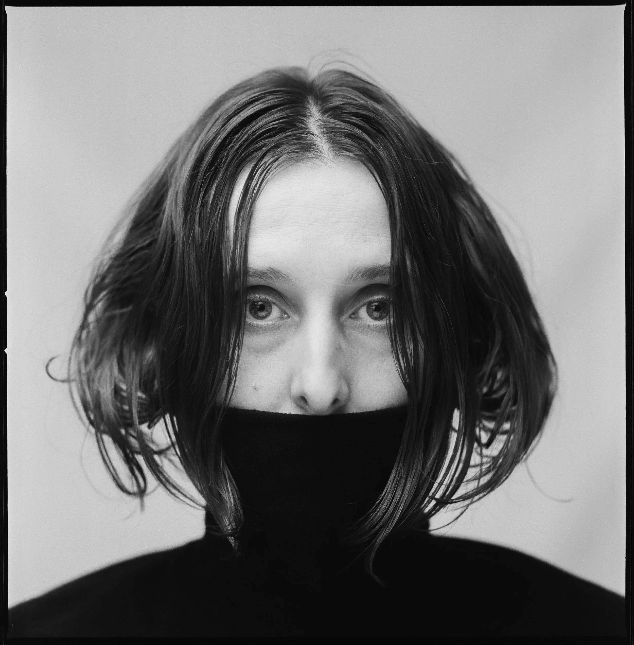 Black and white portrait of a woman with shoulder-length hair, wearing a black turtleneck and a black face mask.