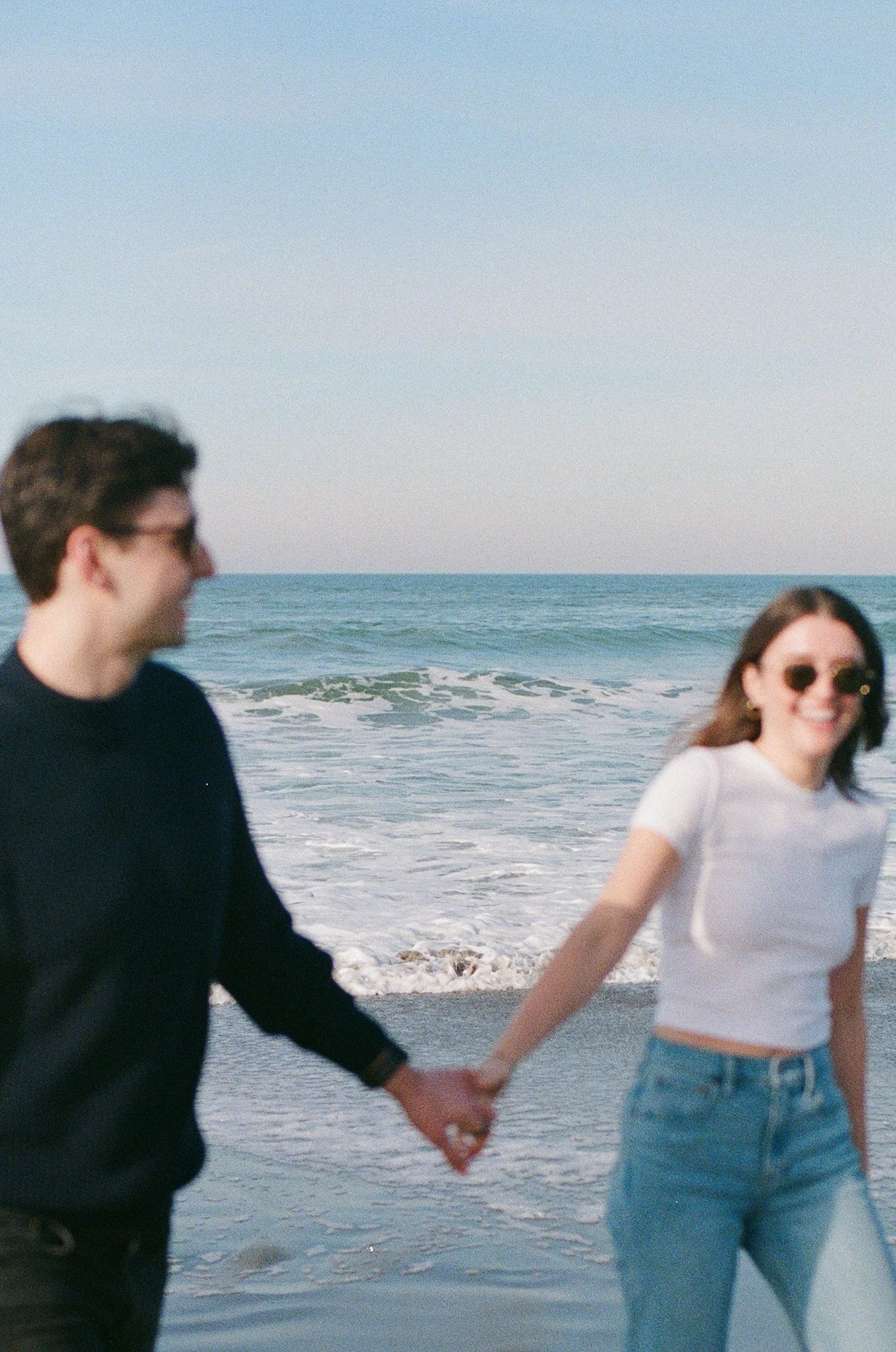 A young man and woman holding hands at the beach, with the ocean and sky in the background. The woman is wearing sunglasses and smiling, dressed in a white t-shirt and jeans, while the man is wearing a black sweater.