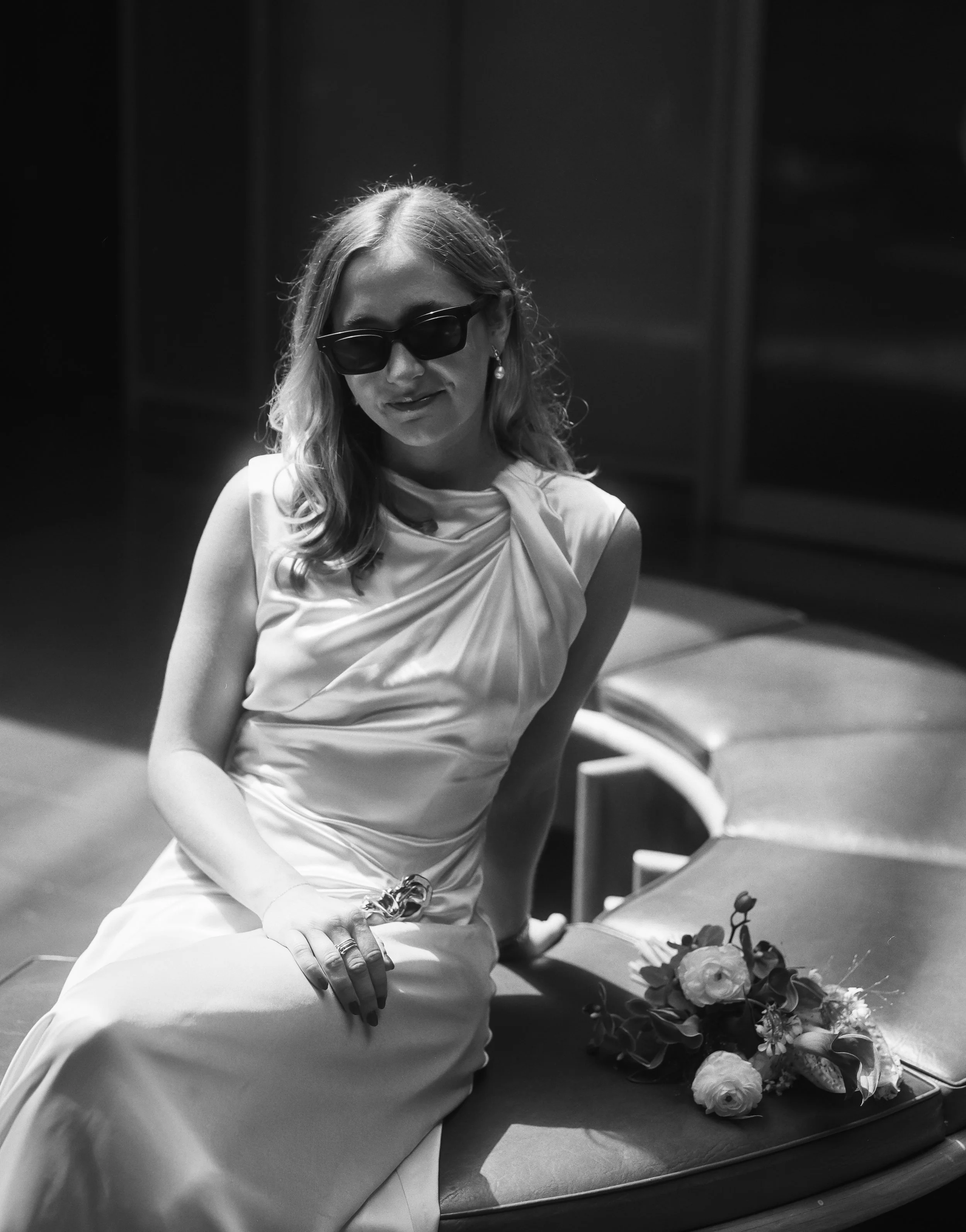 Black and white photo of a woman in a satin dress with sunglasses, sitting on a bench with a bouquet of flowers beside her.