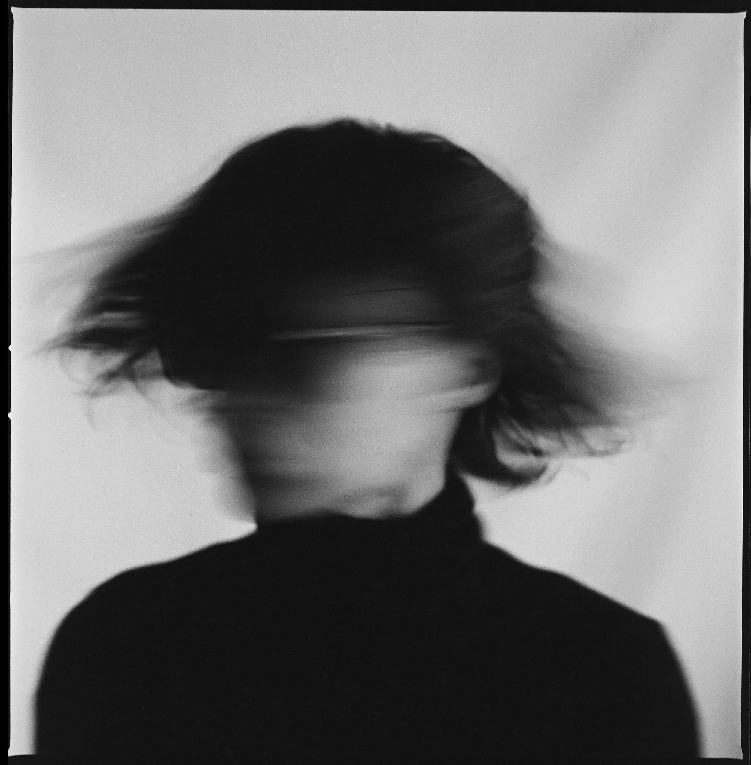 Blurry black-and-white photo of a woman with short hair, head tilted downward, hair swaying to the side, wearing a dark turtleneck, against a plain background.