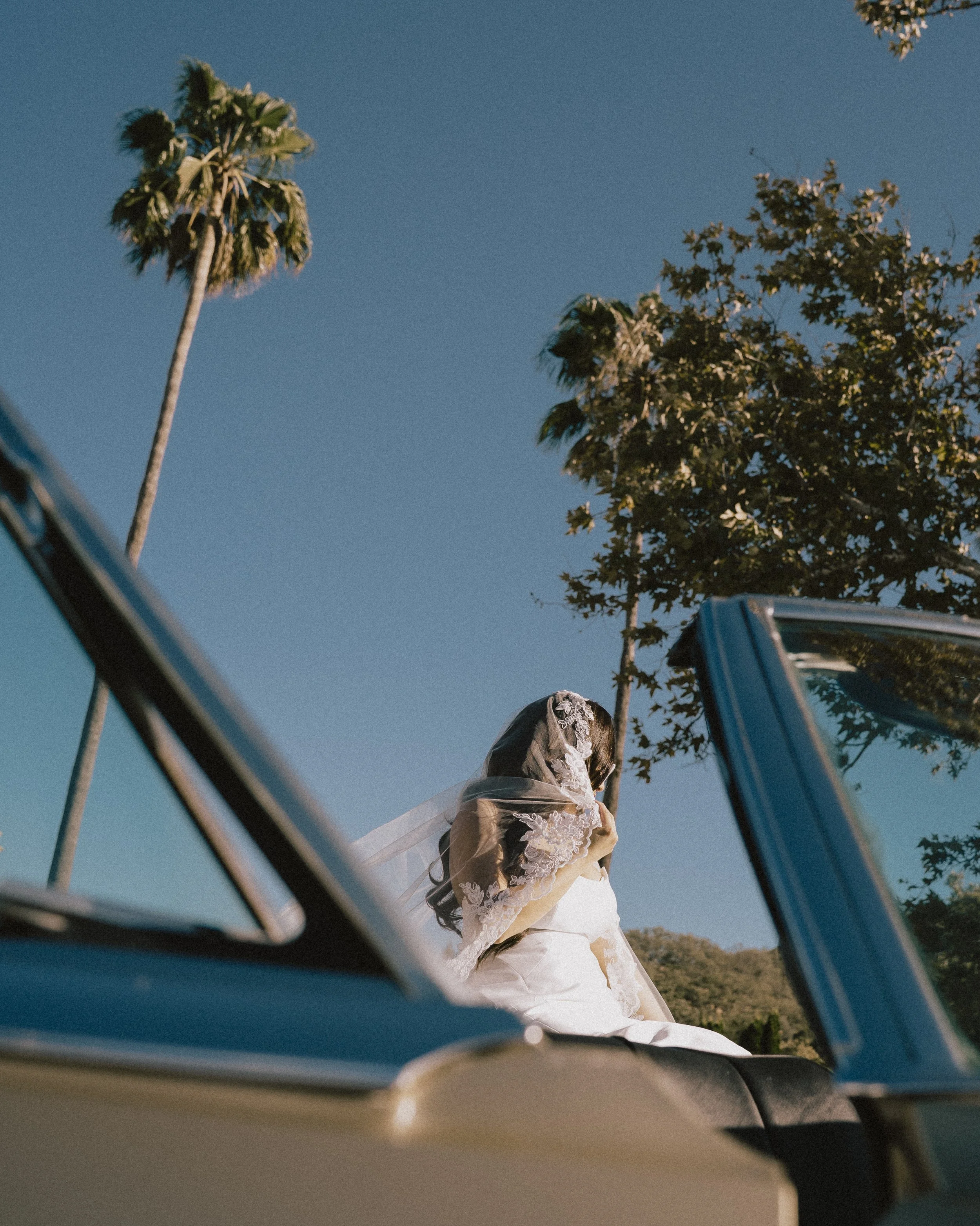 Bride in a white wedding dress and lace veil sitting in a convertible car with open roof, palm trees in background, clear blue sky.