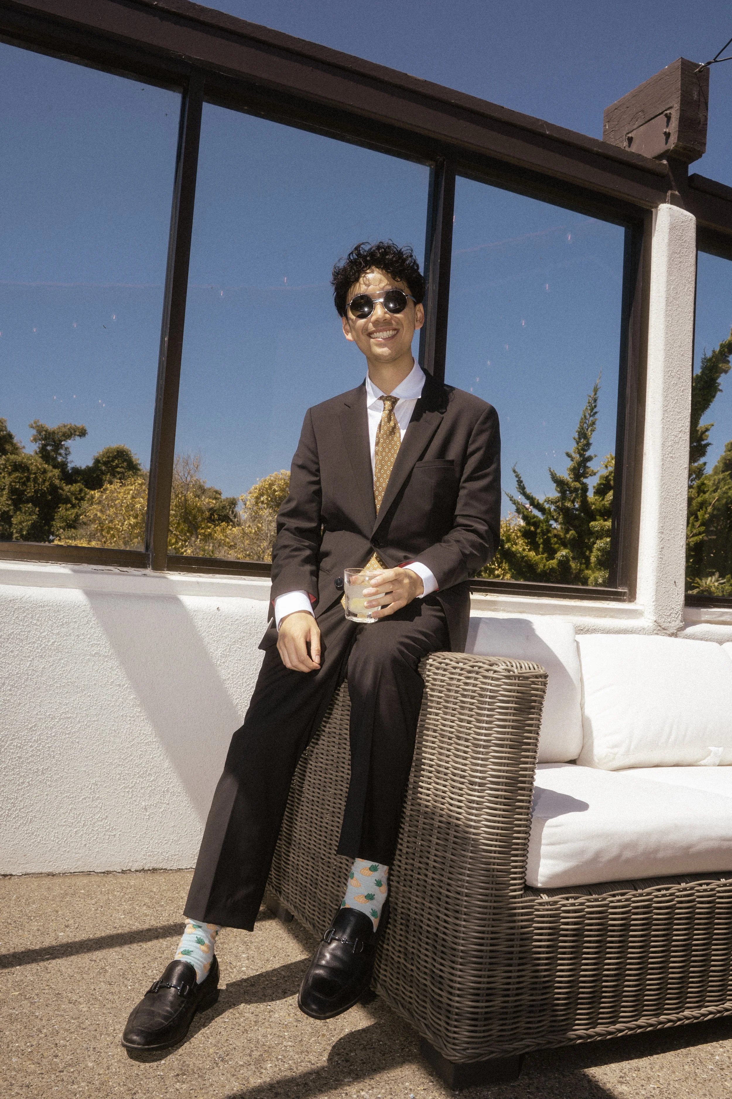 A young man in a black suit, white shirt, and gold tie sits on an outdoor wicker couch holding a glass of drink, smiling at the camera, with trees and a blue sky in the background.