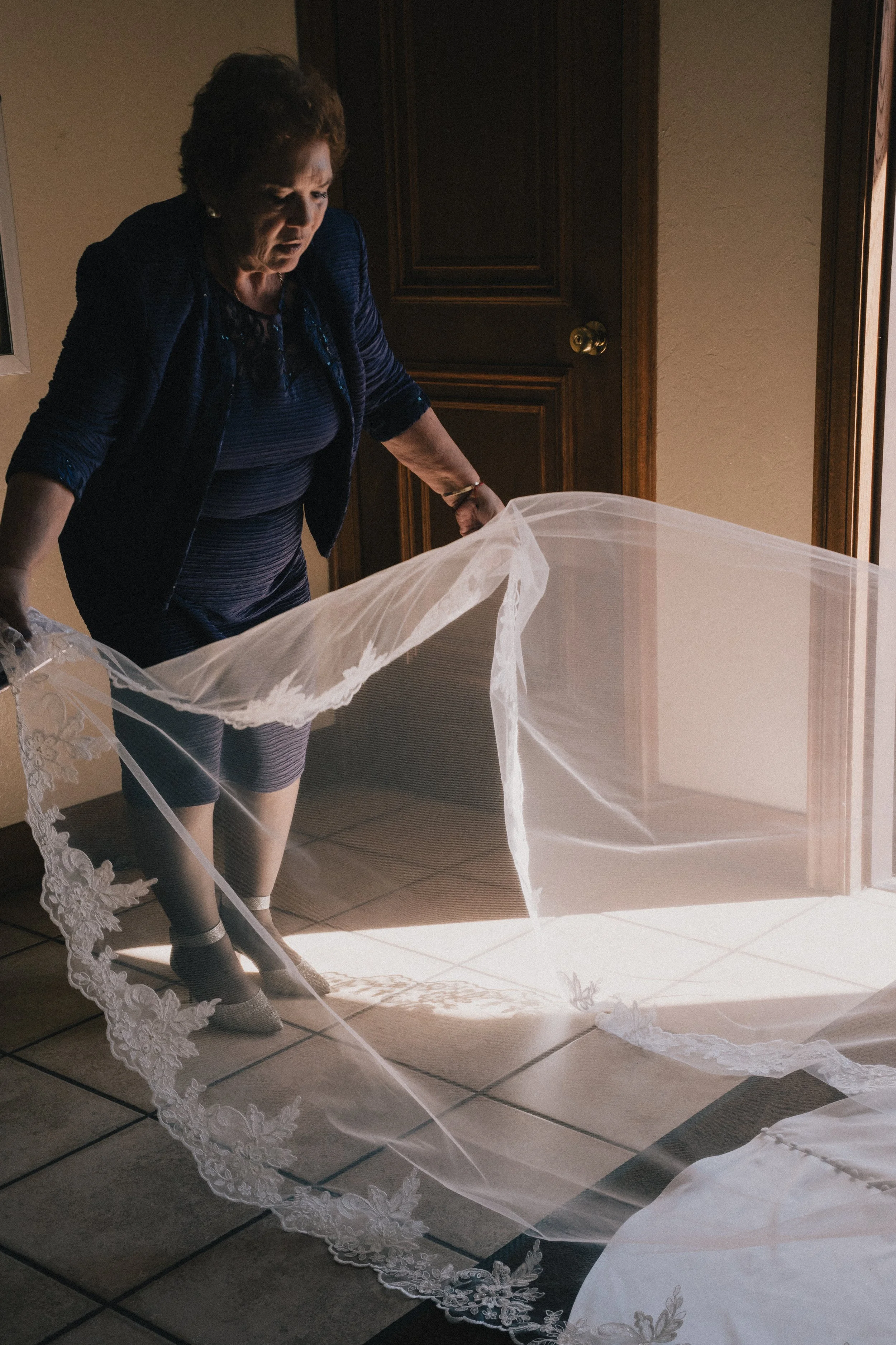 An older woman in a dark blue dress and jacket holding a white lace wedding veil on a tiled floor near a wooden door.