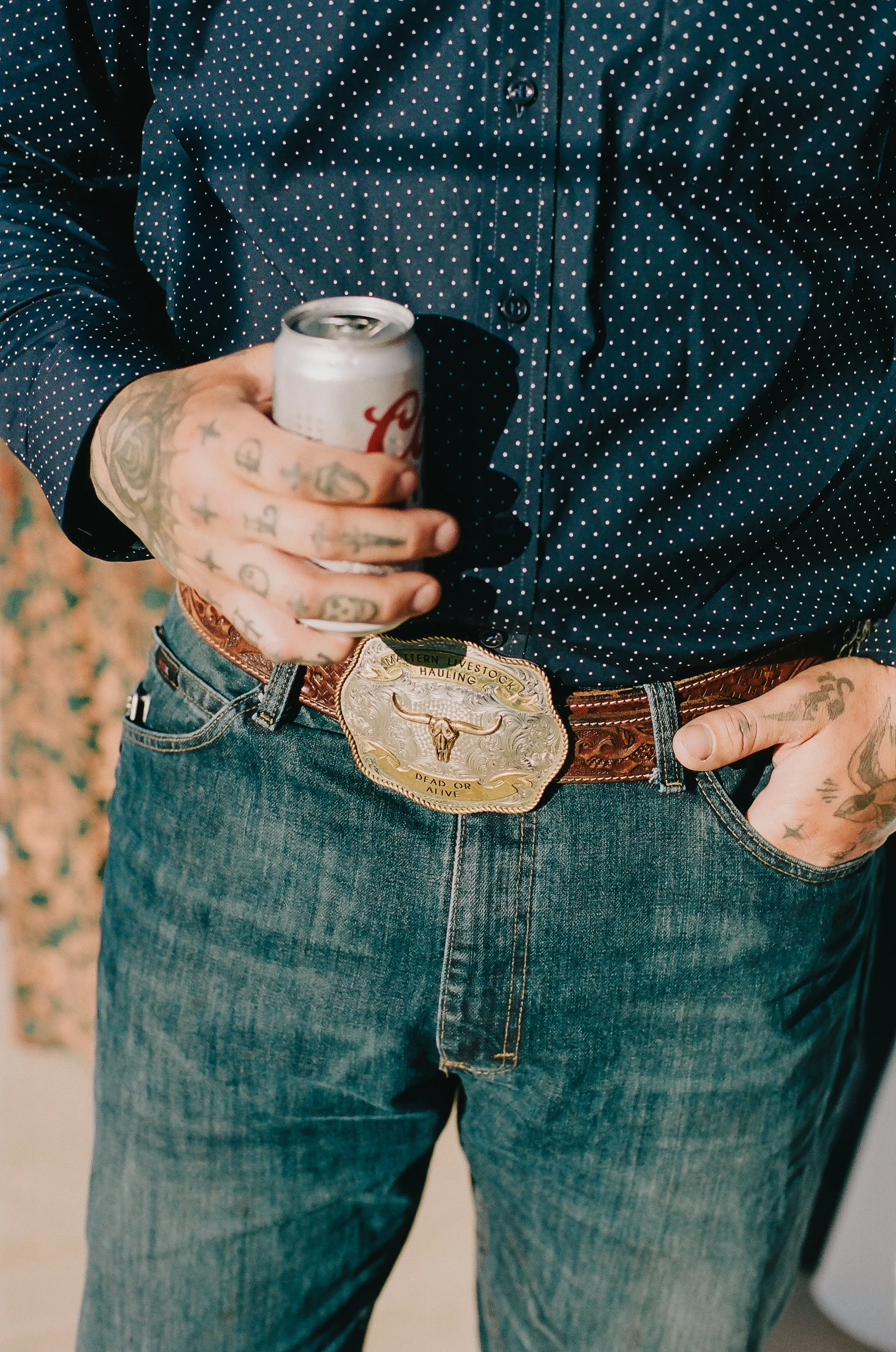 Person wearing blue polka-dot shirt with tattoos on hands, holding a can of soda, with a Western-style belt buckle that reads 'Eastern Livestock Hauling, Dead or Alive' and jeans.