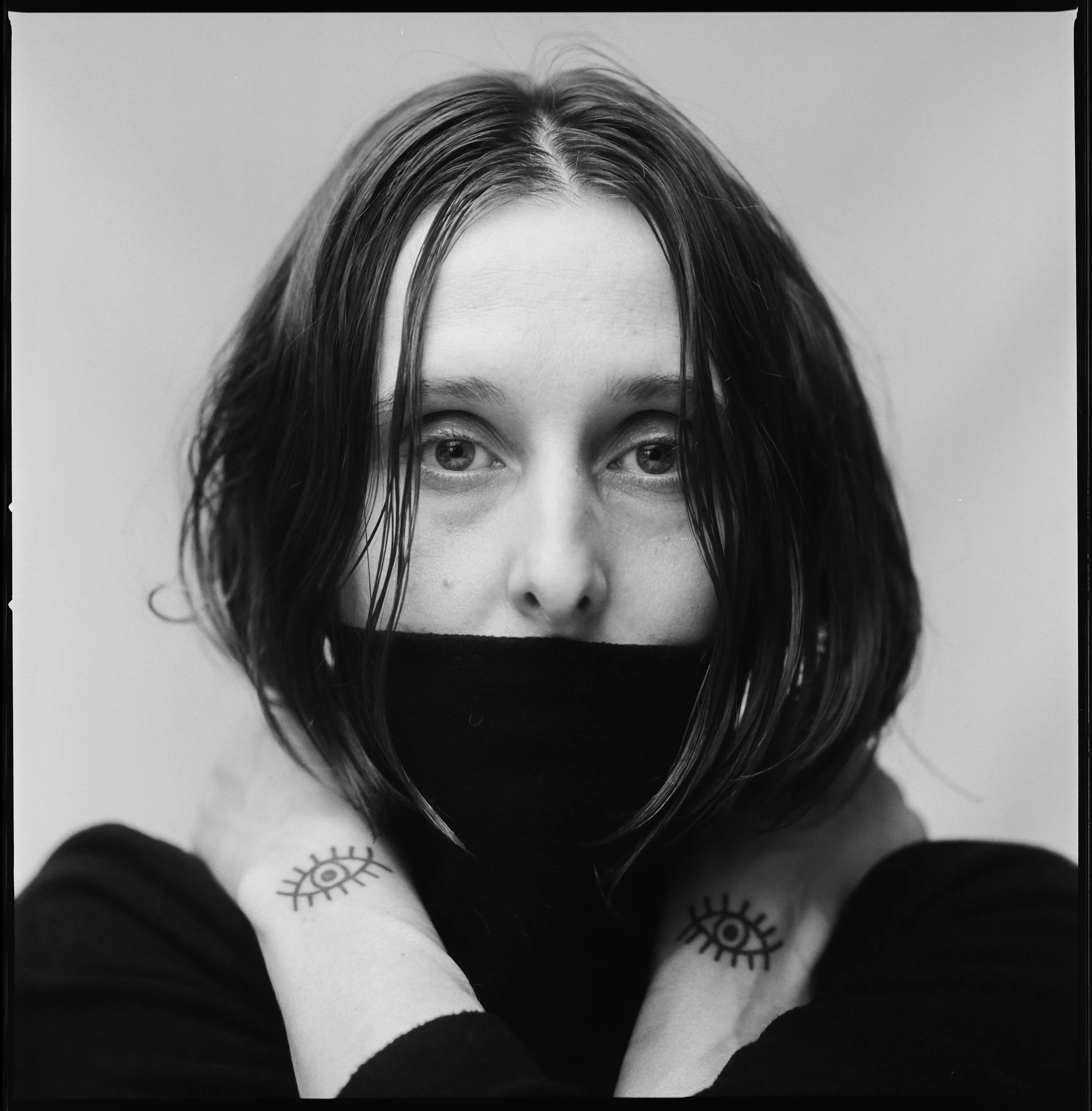 A black-and-white portrait of a woman with short, wet-looking hair, wearing a face mask, holding her neck with both hands, and showing tattoos of eyes on her wrists.
