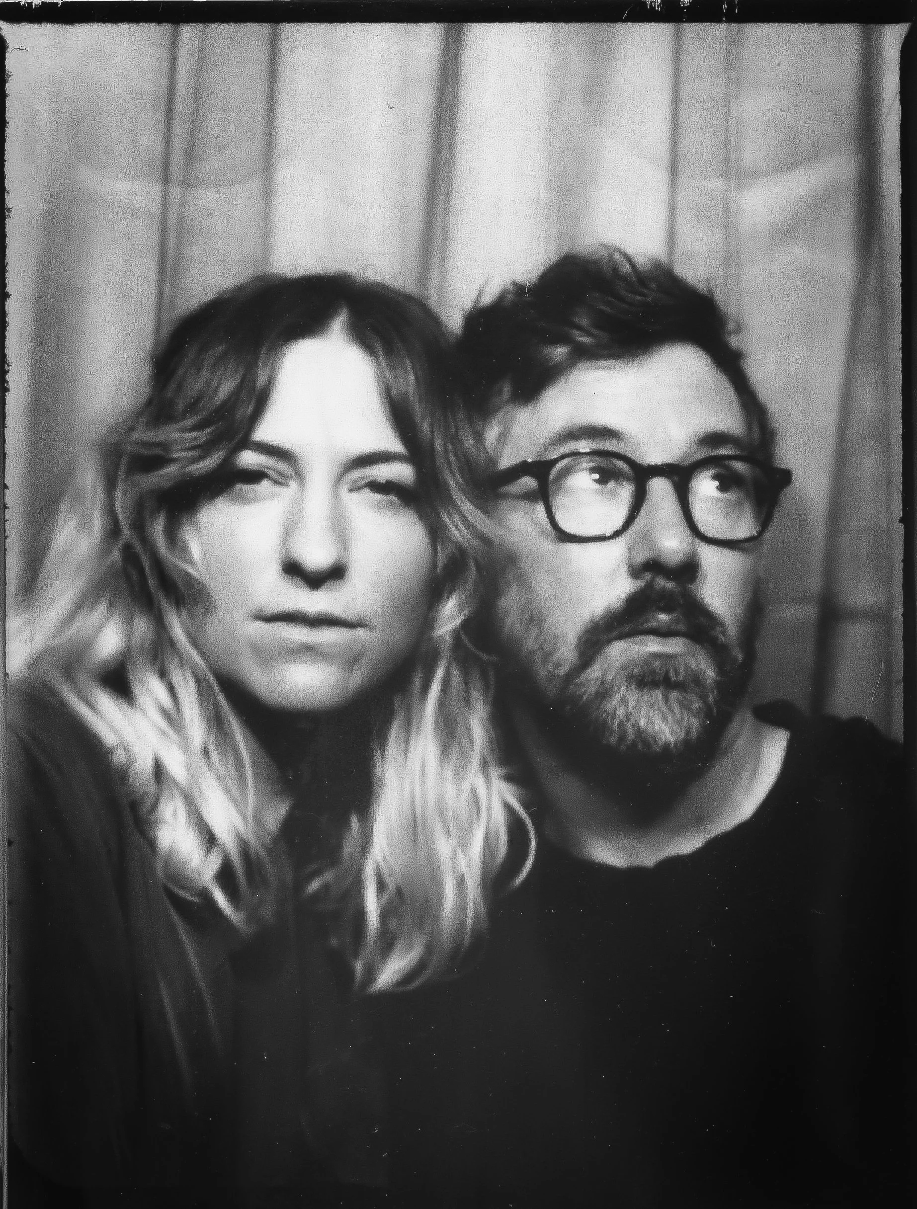 Black and white photo of a woman with wavy hair and a man with glasses and a beard, both looking serious, standing in front of curtain background.