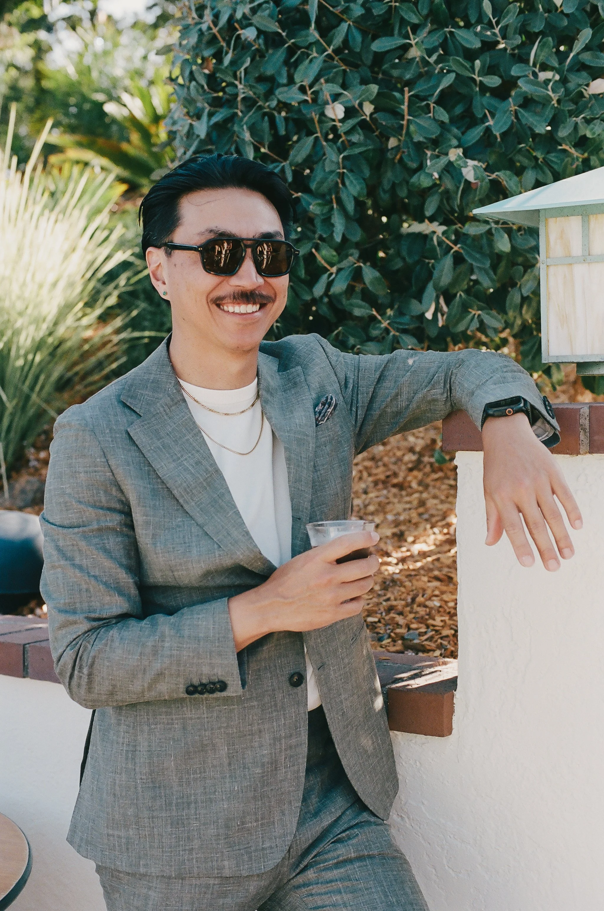 A man in a gray suit with black sunglasses, a mustache, and earrings, smiling while holding a drink outdoors near a white wall and lush greenery.