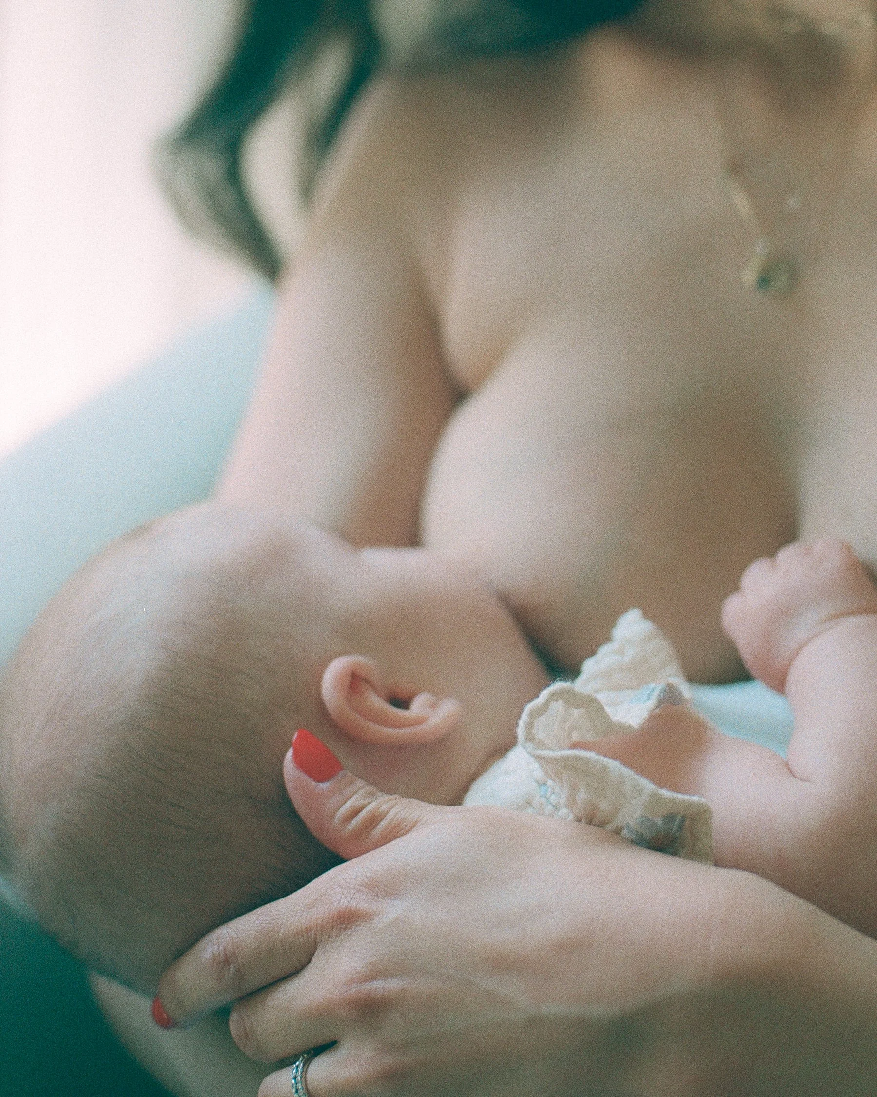 A woman holding a sleeping newborn baby close to her chest, with her hand supporting the baby's head. The woman is wearing a necklace and has red nail polish.