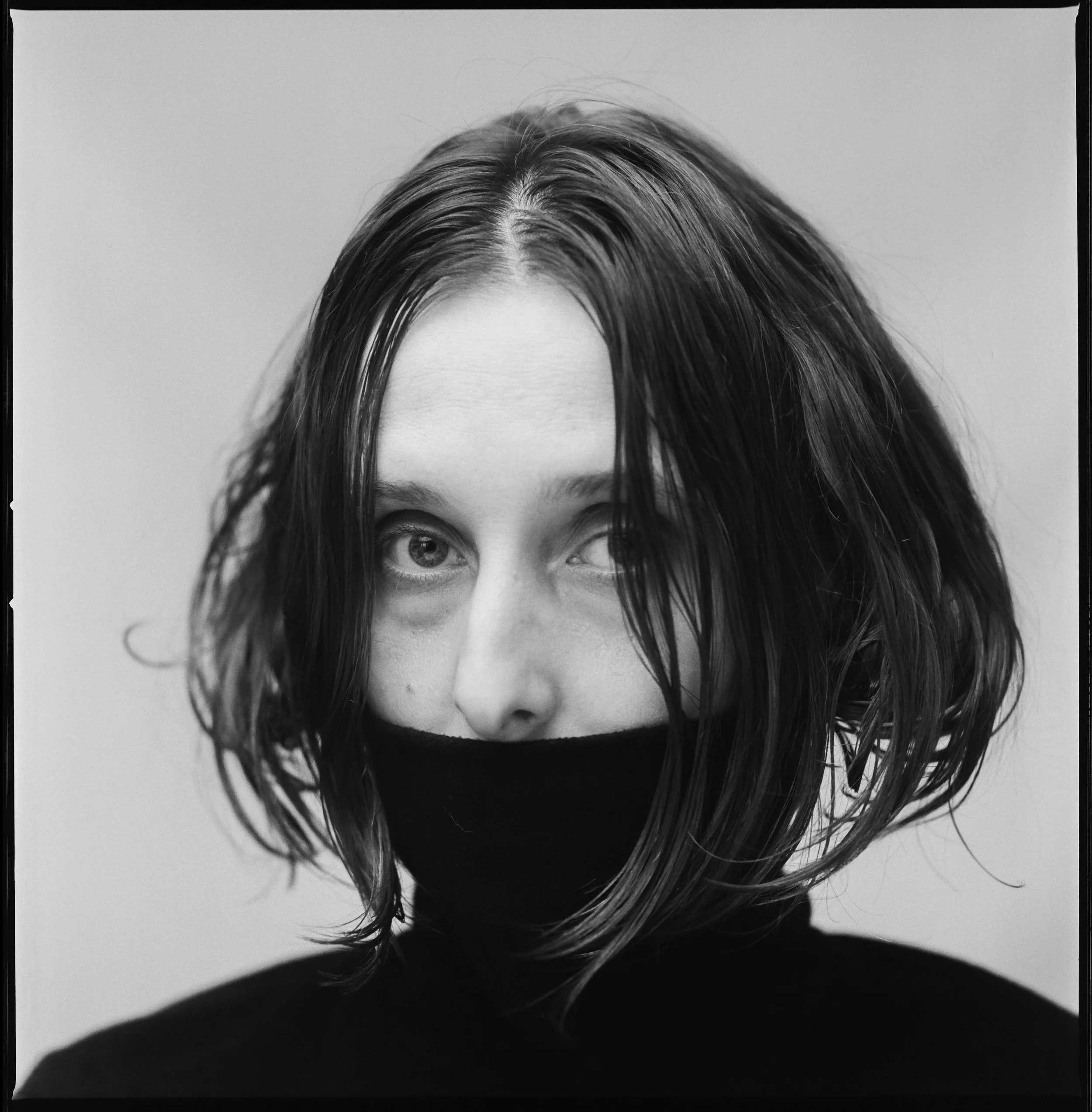 Black and white portrait of a woman with wavy hair, wearing a black face mask and a black turtleneck, with one eye winking.