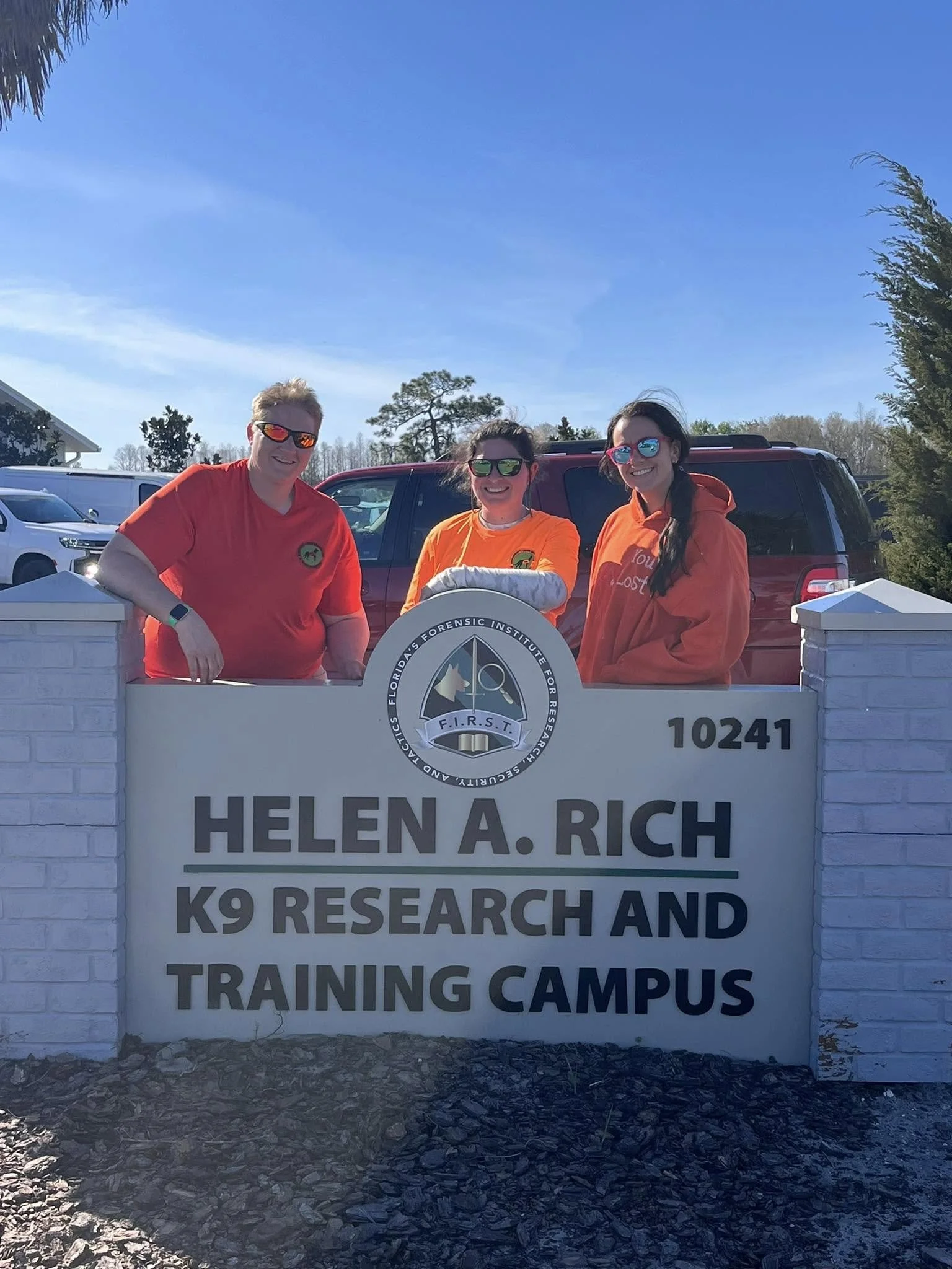 Three women standing behind a white brick sign with a logo and text that reads 'Helen A. Rich, K9 Research and Training Campus' at a research facility. They are outdoors with cars and trees in the background, wearing sunglasses and orange shirts.