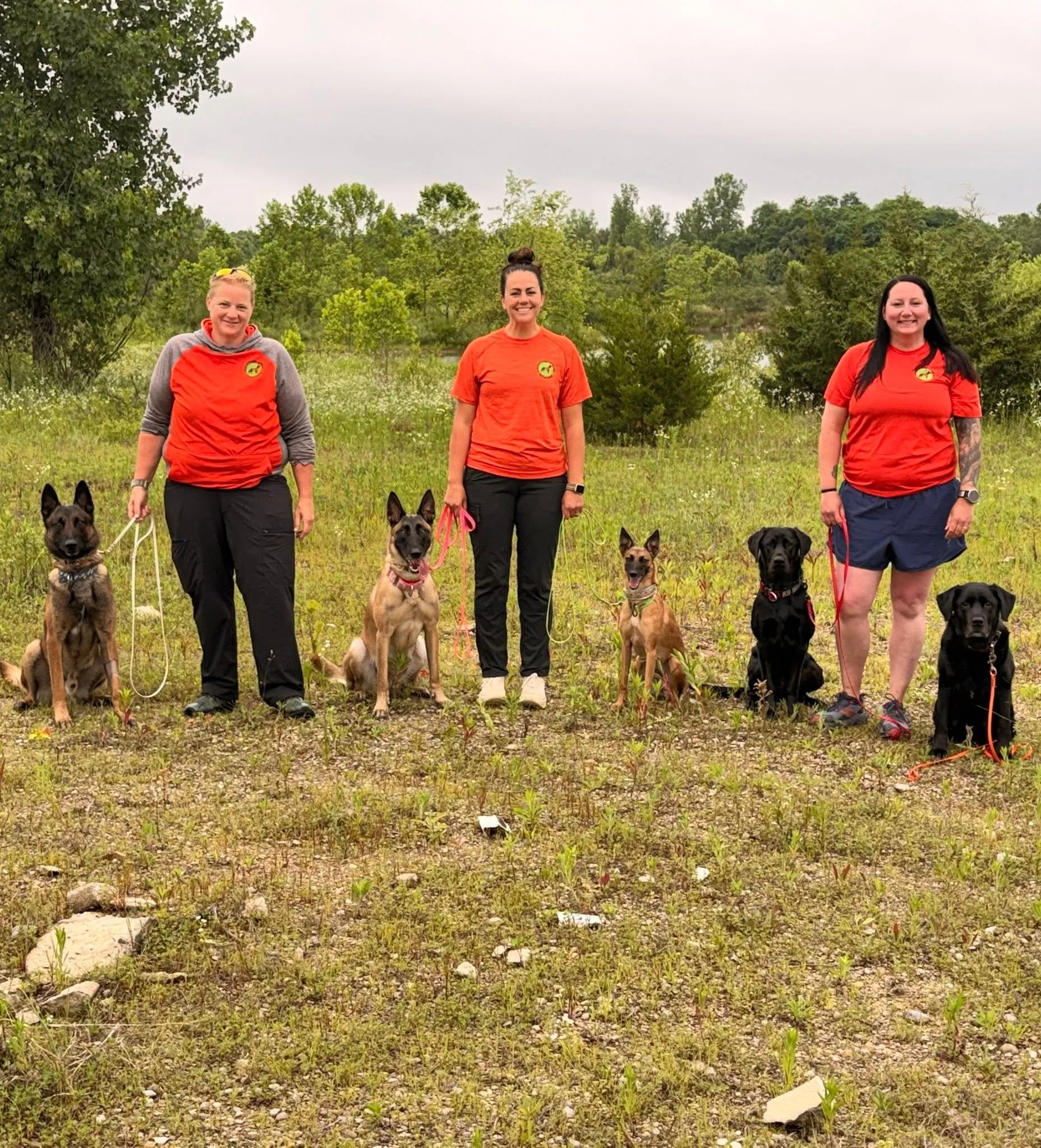 Three women standing outdoors with five dogs in a grassy area surrounded by trees, under an overcast sky.