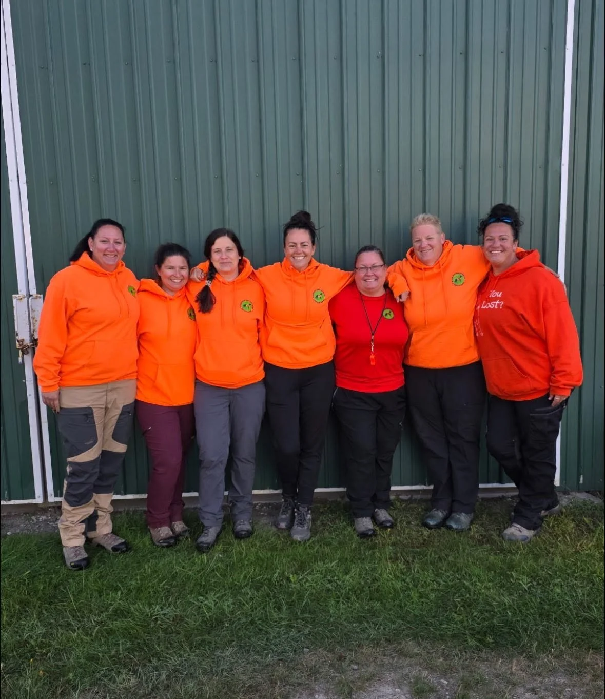 Group of seven women standing outdoors in front of a green metal building, smiling, wearing orange hoodies and outdoor gear.