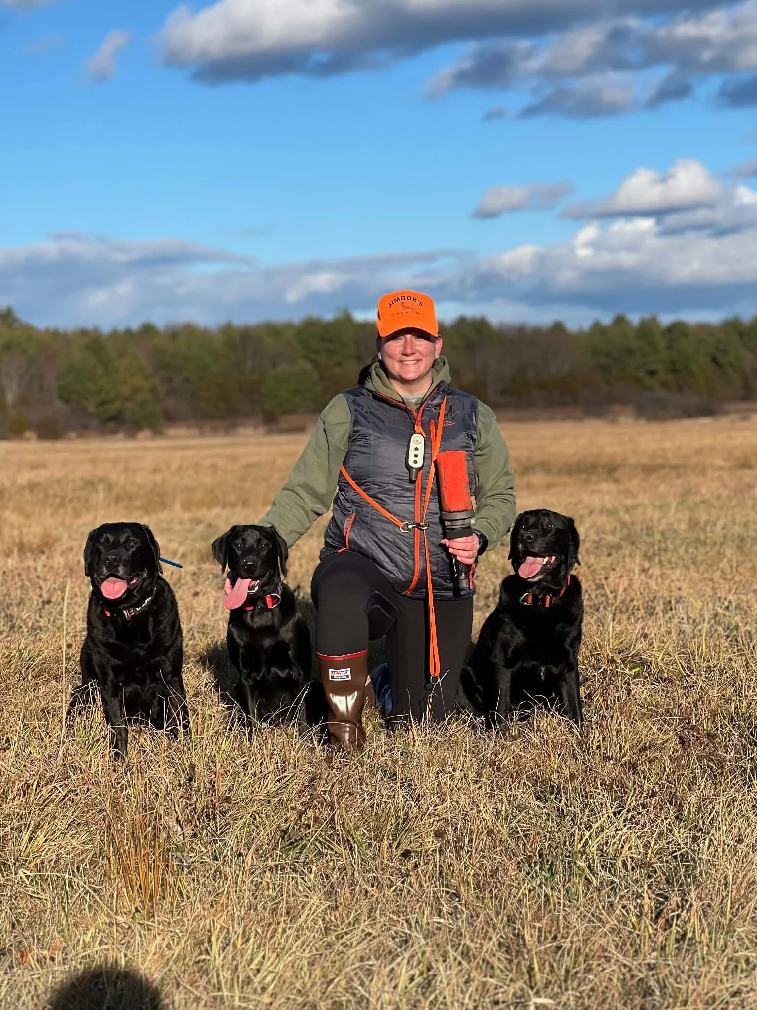 A woman standing in a field with four black Labrador retrievers, wearing a bright orange cap and outdoor gear, with a background of trees and a sky with clouds.