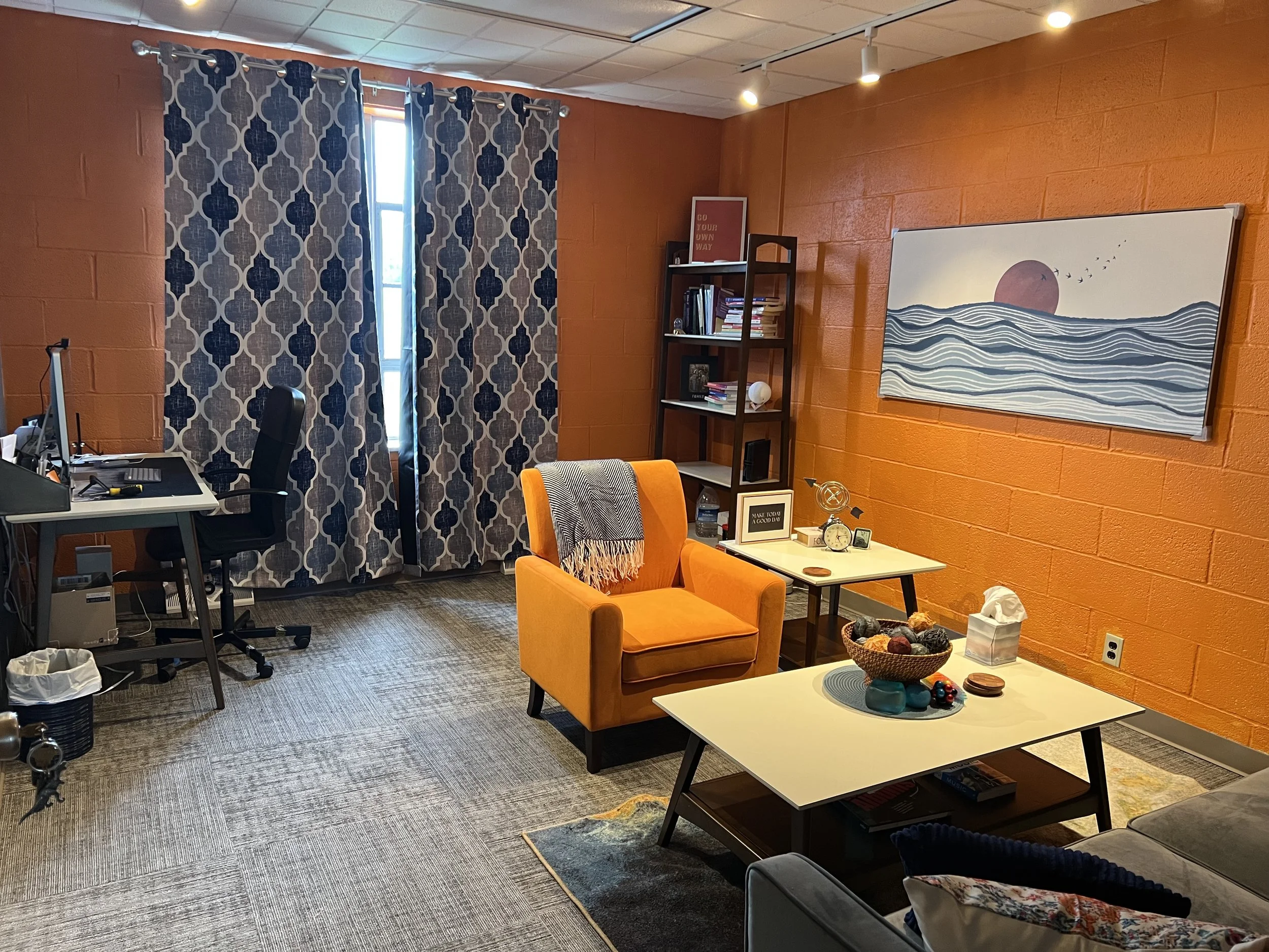 A cozy home office and living area with a black desk and chair, an orange armchair with a throw blanket, a white coffee table with decorative items, a bookshelf, a wall painting of wavy water and a sunset, and patterned curtains on a window, with warm orange brick walls.