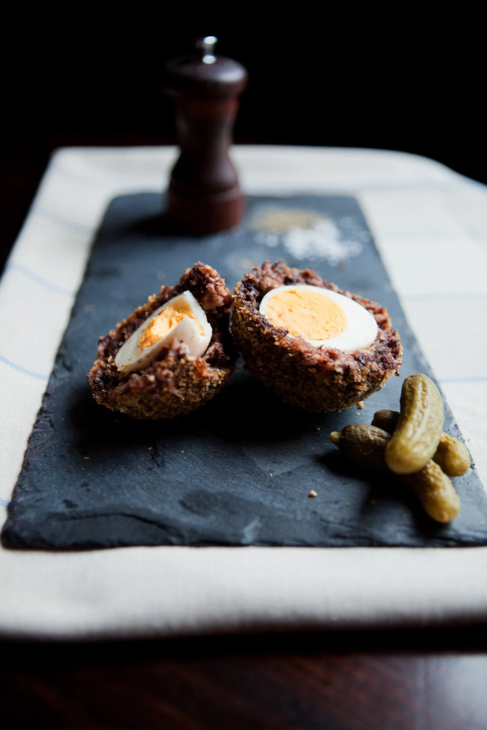A slice of Scotch egg with chopped egg in a crunchy breaded exterior on a black slate serving board, with a small pile of salted cashew nuts beside it. In the background, a pepper mill is visible. Image by Celine Marchbank.