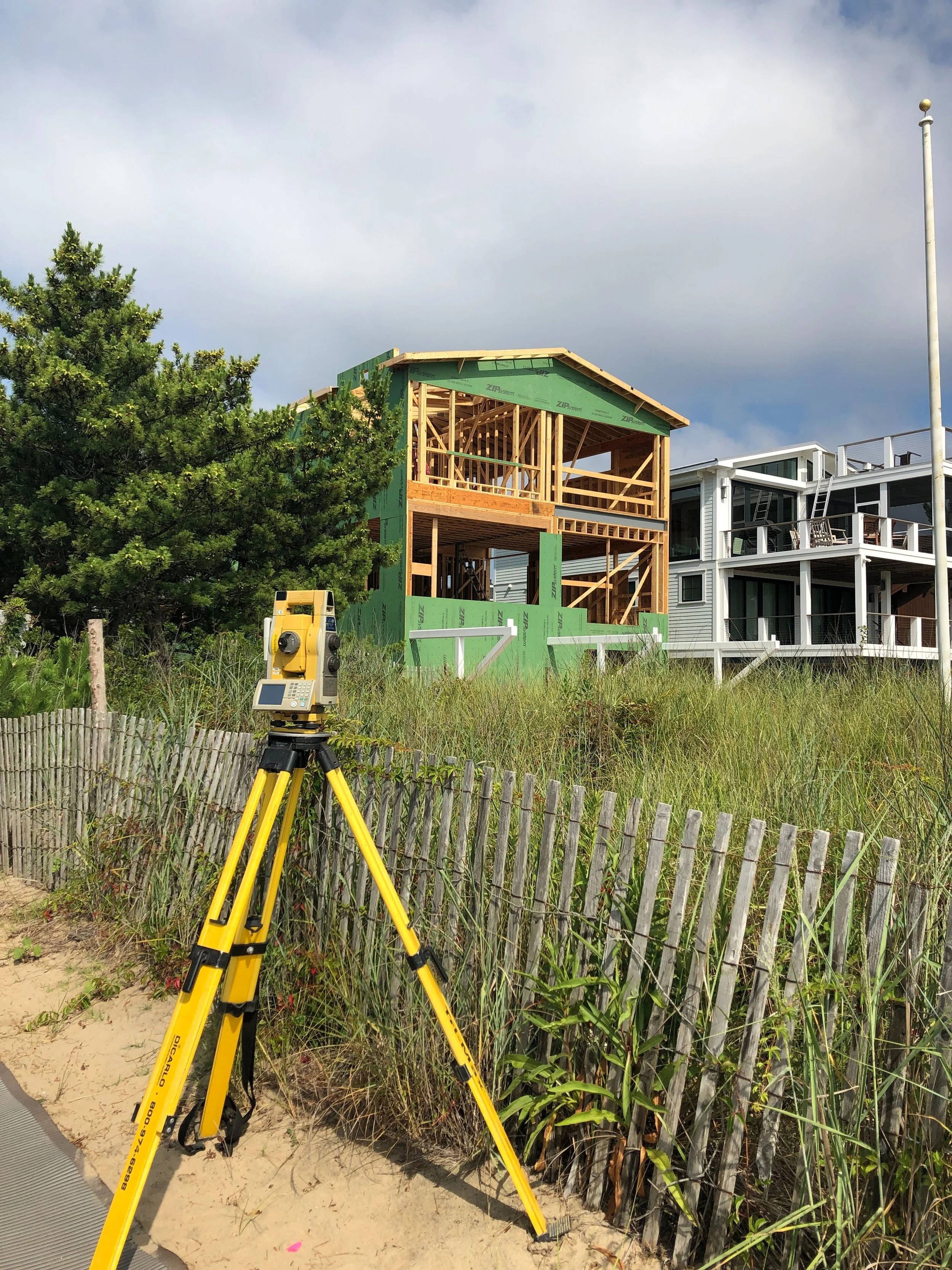 Construction site with a partially built wooden house, a surveying instrument on a tripod, grassy sand dunes, and a neighboring modern white house with balconies.
