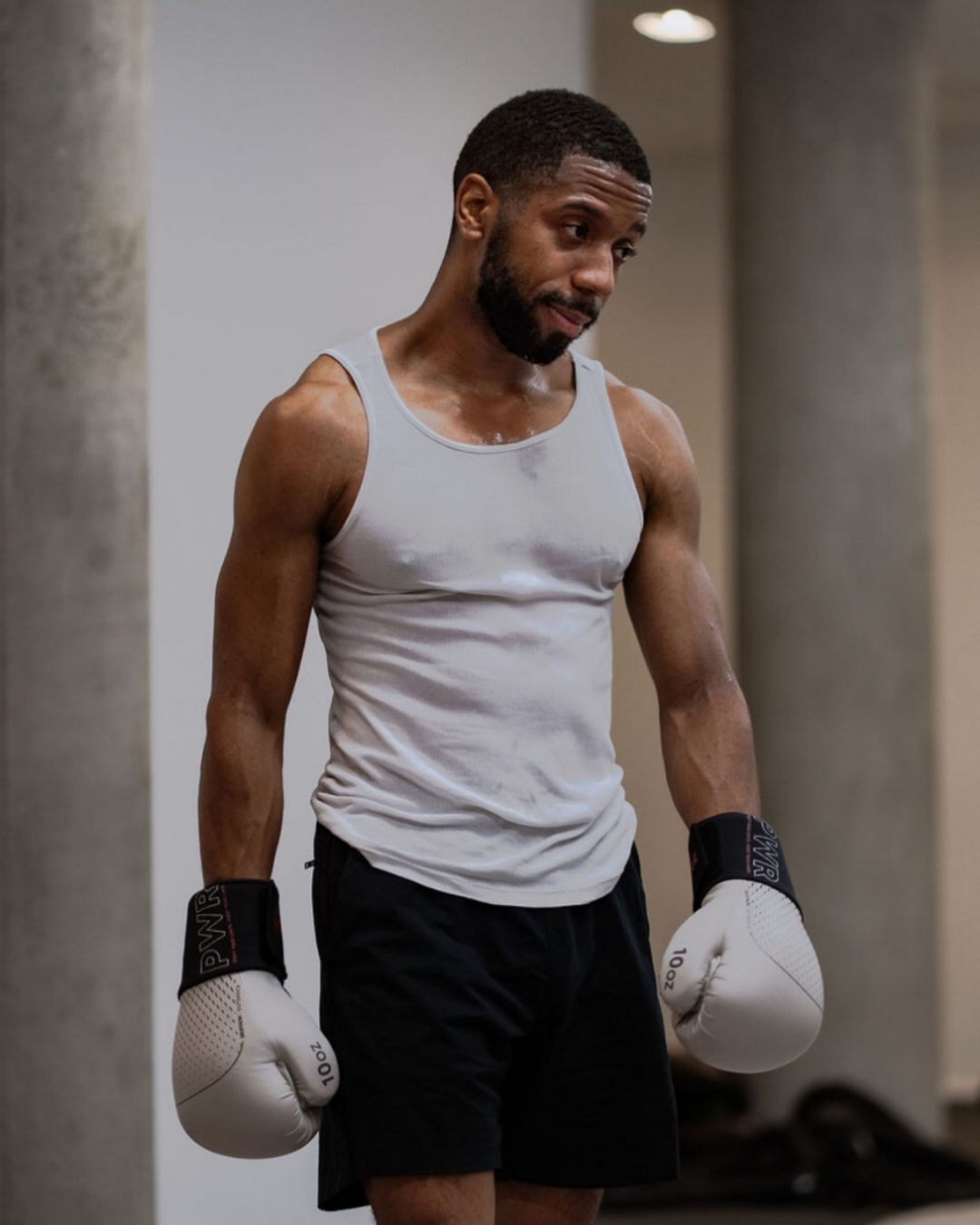 A man wearing a white tank top and black shorts is standing in a gym with boxing gloves on, looking down with a contemplative expression.