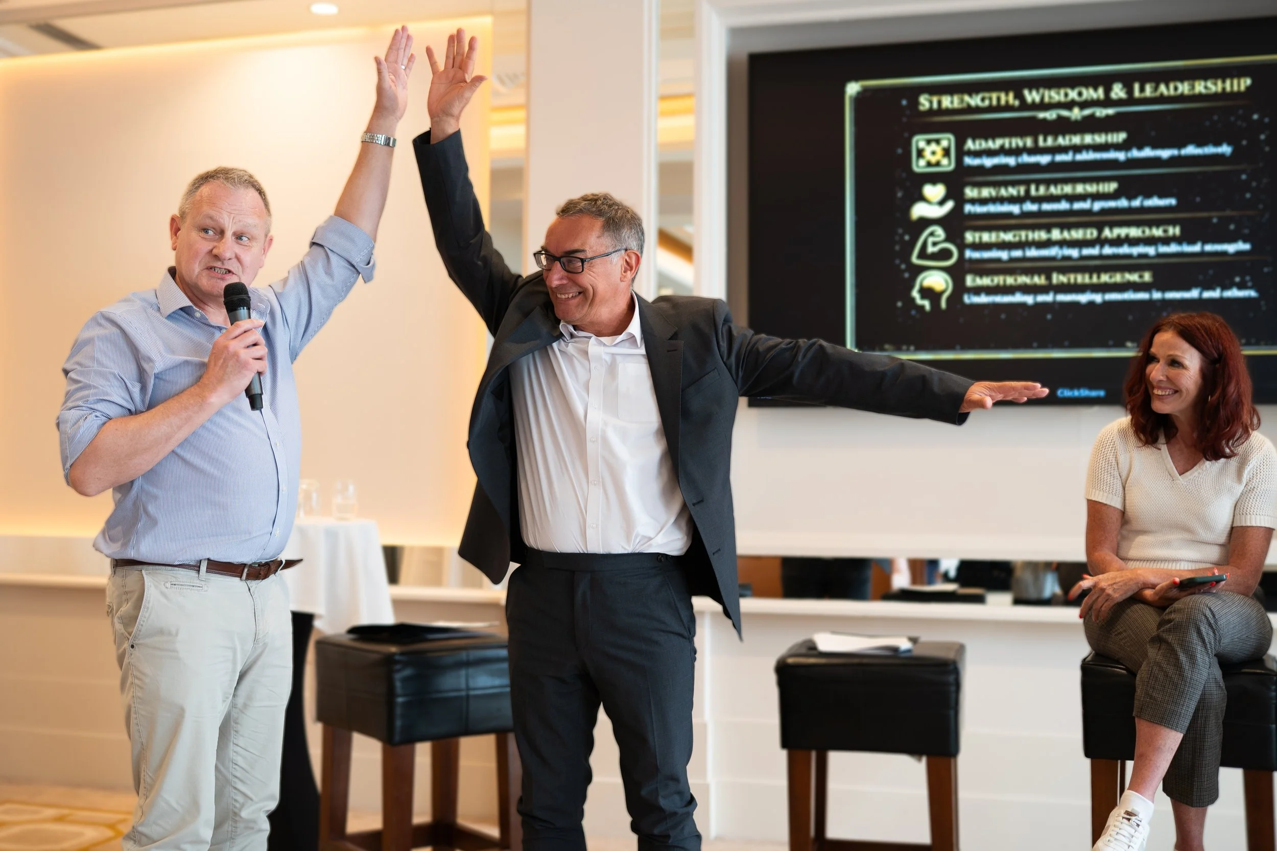 Two men, one holding a microphone, raising their hands together in a gesture of celebration or agreement during a presentation or conference. A woman is seated nearby, smiling and holding a phone. There is a large screen in the background displaying 