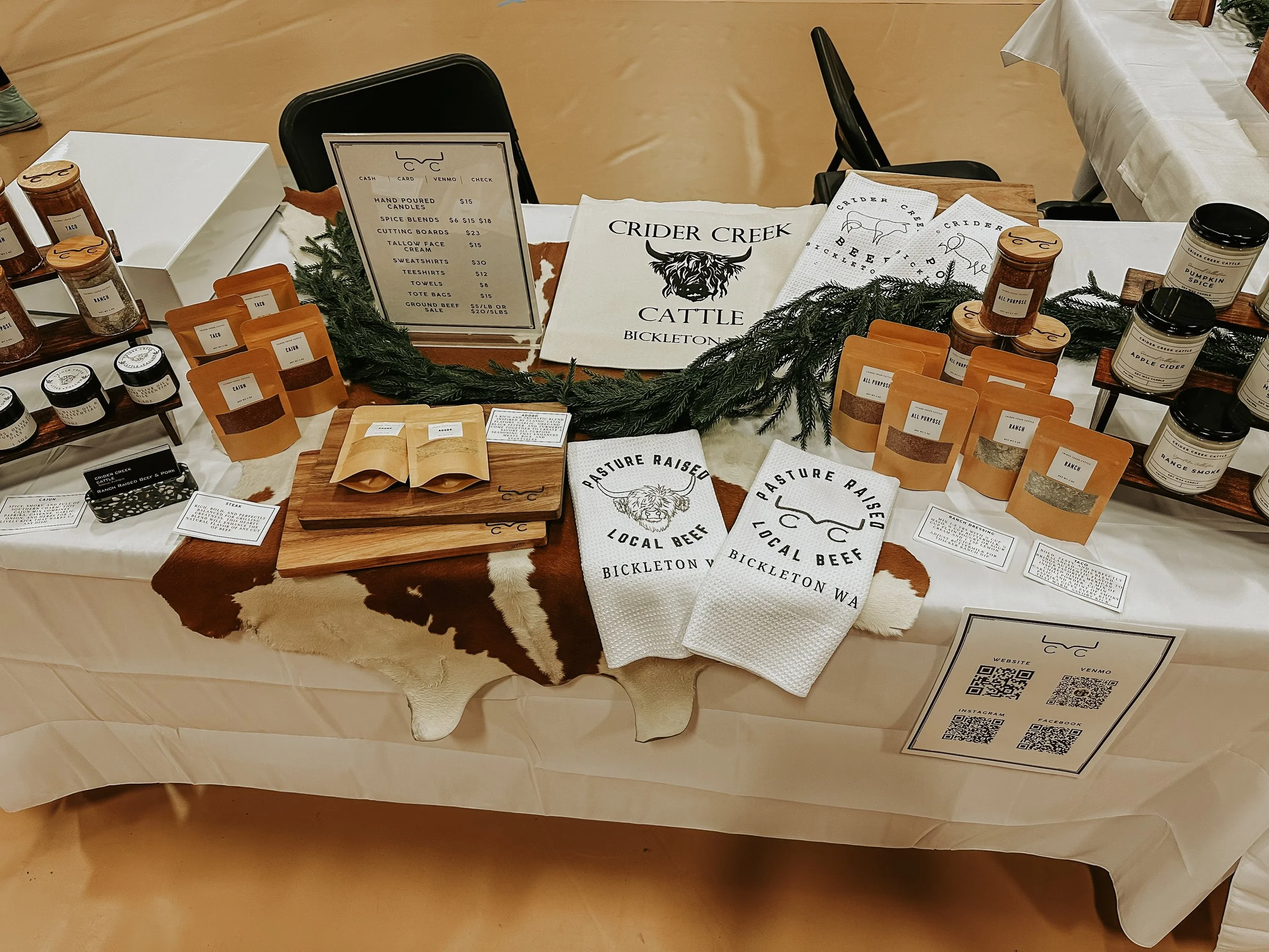 Market stall display with jars, packets, and signs promoting Crider Creek cattle and local beef, decorated with evergreen branches, on a long table with a cowhide table runner.