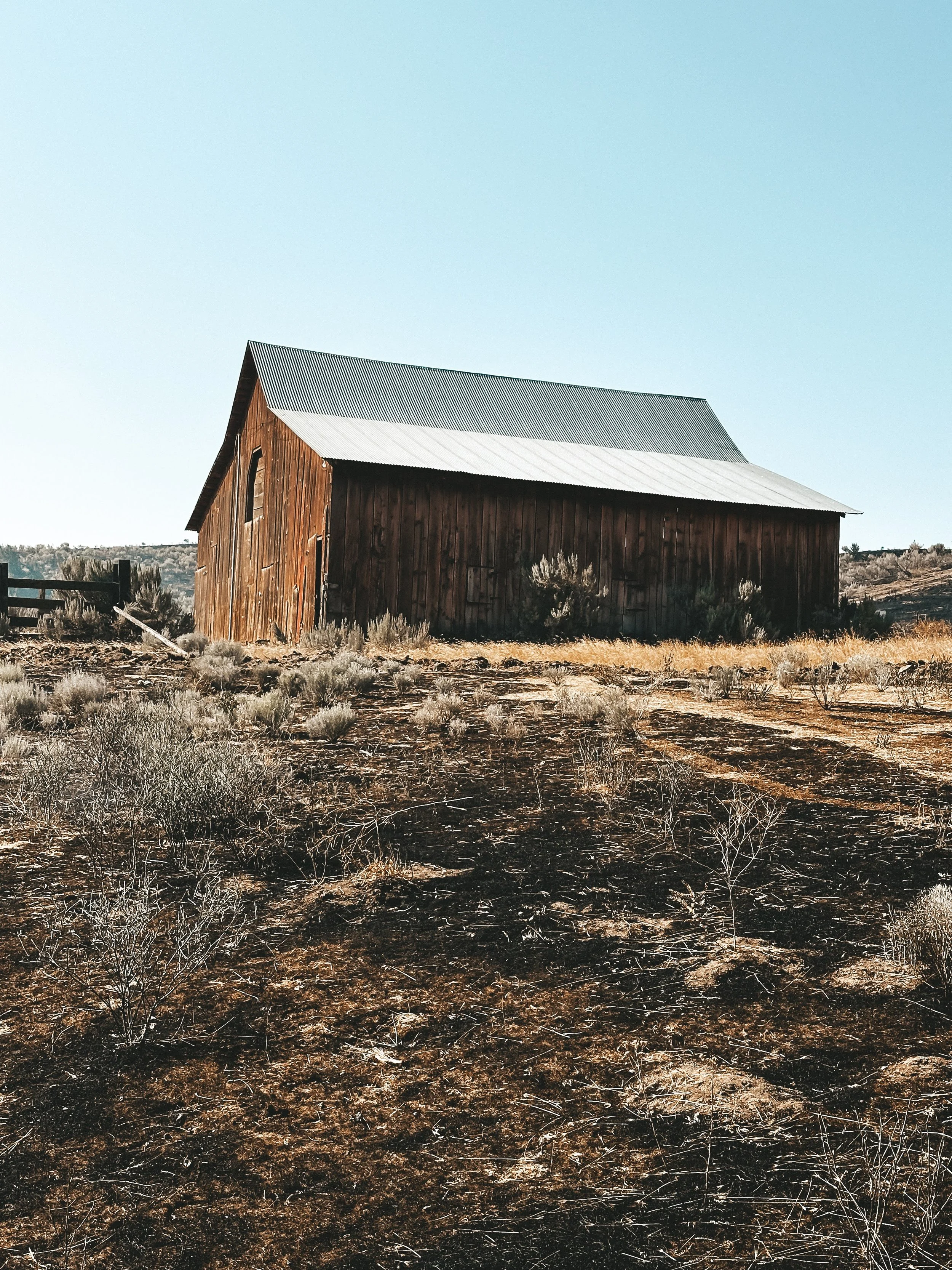 A rustic wooden barn with a silver metal roof situated in a semi-arid landscape with dry grass and sparse shrubs under a clear blue sky.