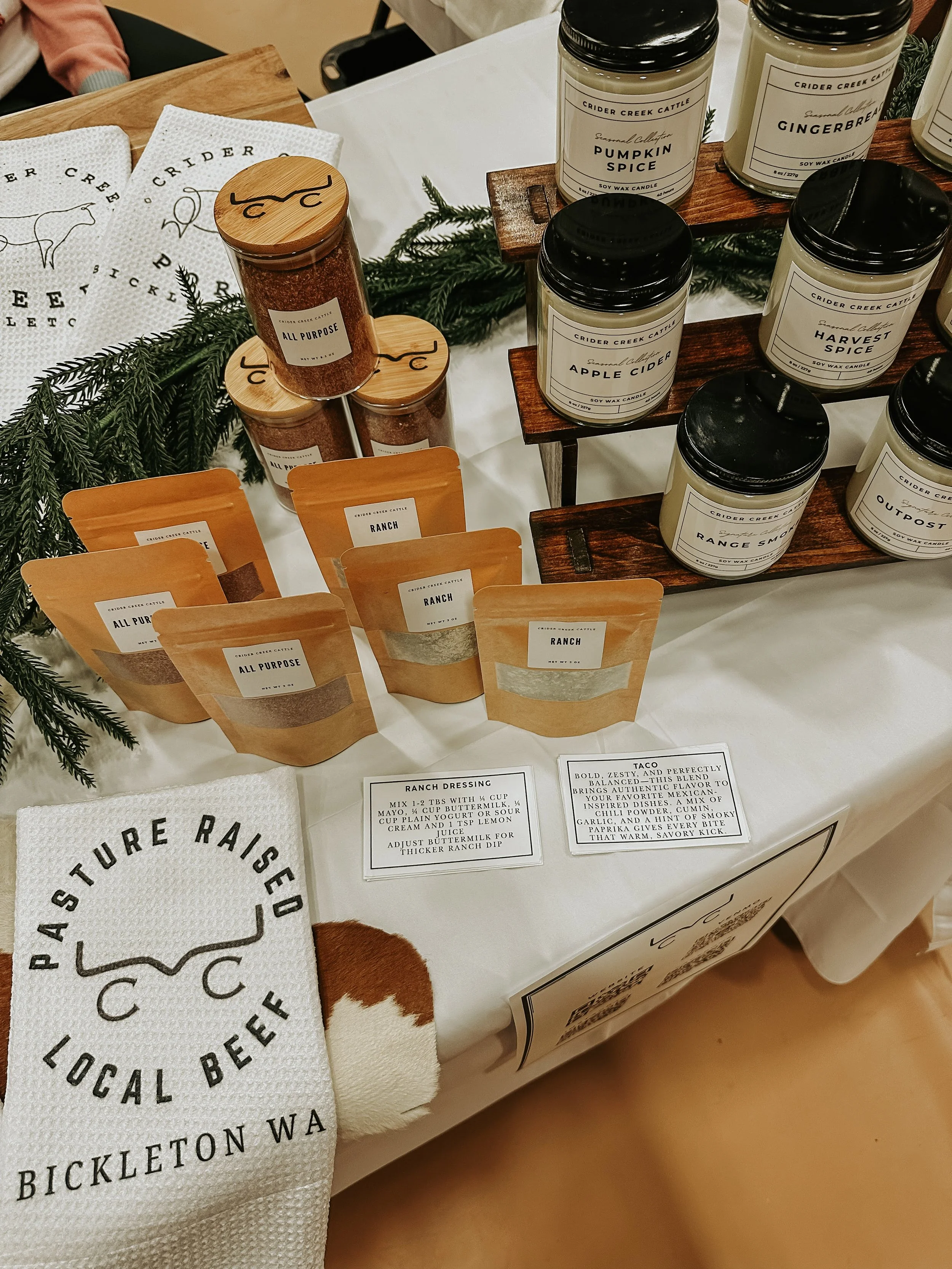 Display of various scented candles, spice mixes, and seasonings on a table at a local beef event in Bickleton, Washington. Includes jars labeled pumpkin spice, gingerbread, apple cider, harvest spice, range smoke, and outpost, as well as seasonings labeled all purpose and ranch, with recipes and informational cards.
