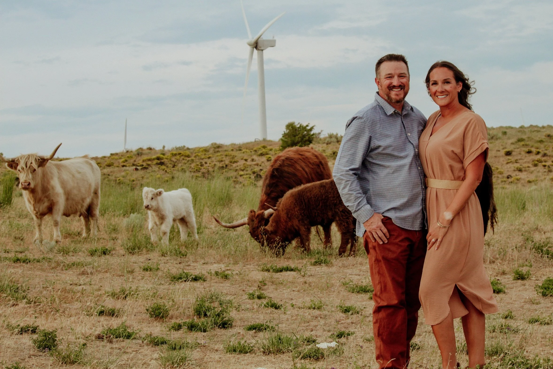 A smiling couple stands together in a rural field with grazing cattle and wind turbines in the background.