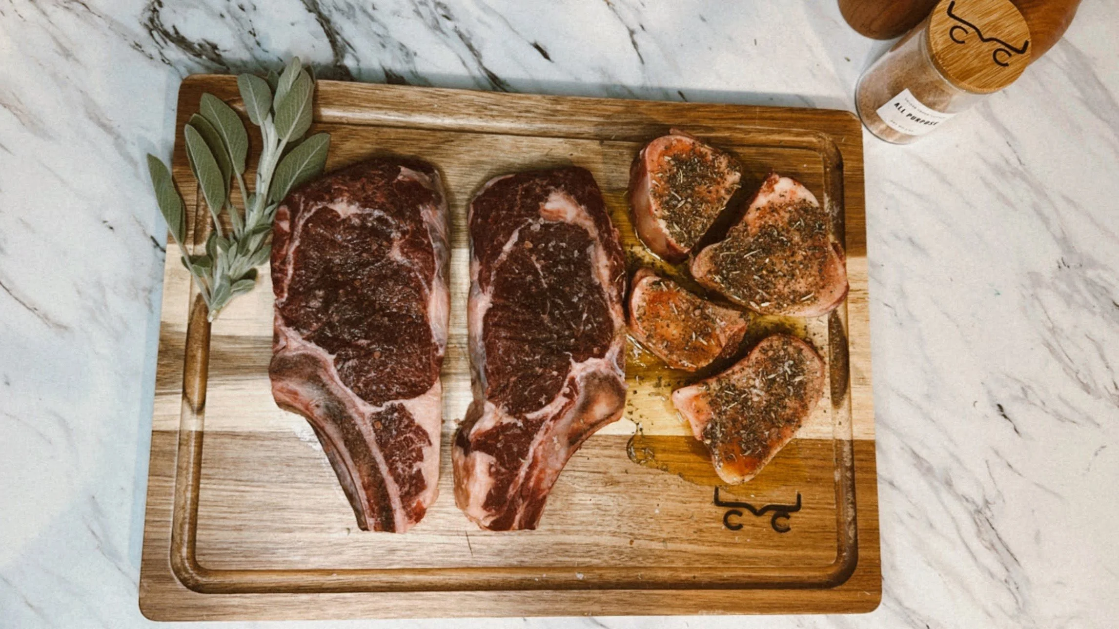 Two raw beef steaks and four seasoned tuna steaks on a wooden cutting board, with a sprig of sage and a glass jar of spices nearby.