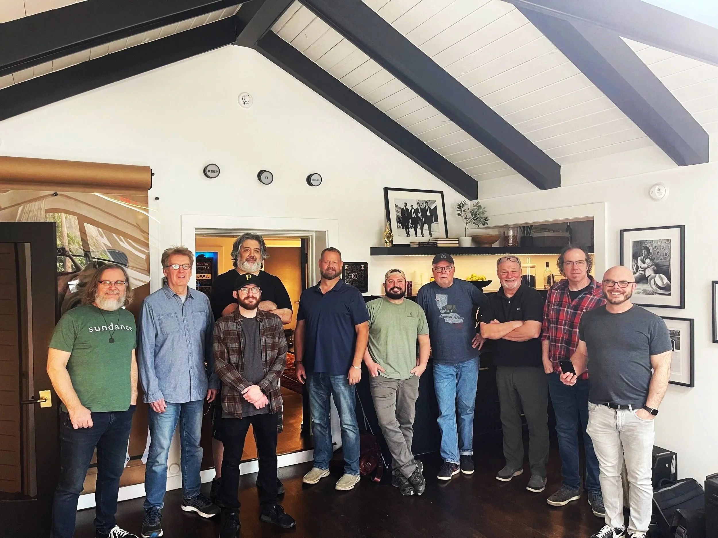 Group of eleven men standing inside a Nashville recording studio lounge area, casually posed in a horizontal lineup. The room features framed photos on the wall, studio gear visible in the background, and warm interior lighting. The men appear relaxe
