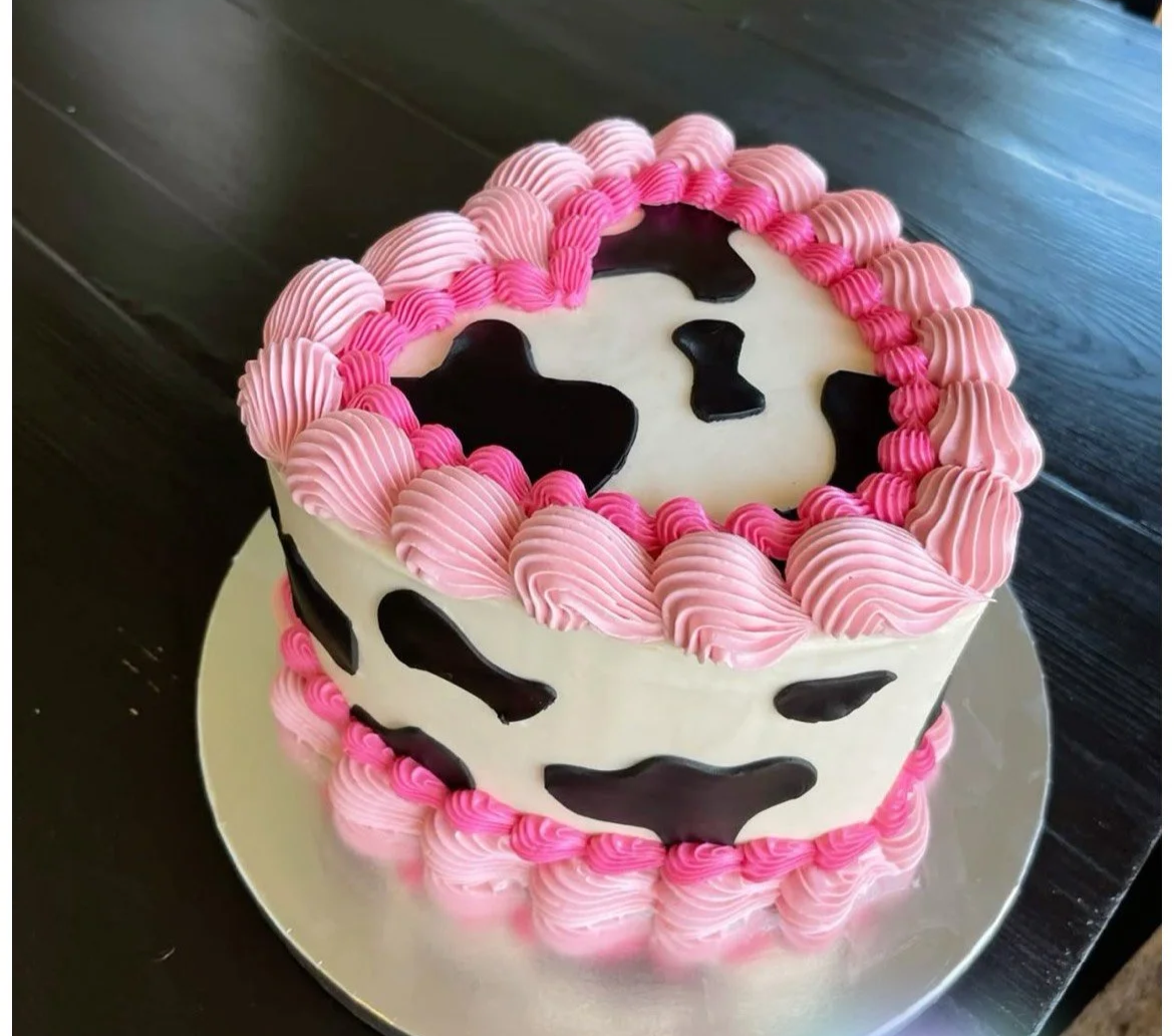 Group of women and men decorating cakes and flowers at a pink-draped table in a room, with a large TV mounted on the wall.