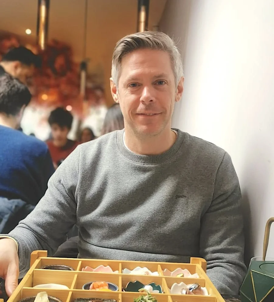 Andy Windsor - Director. A man sitting at a table in a restaurant with a box of sushi in front of him, smiling at the camera.