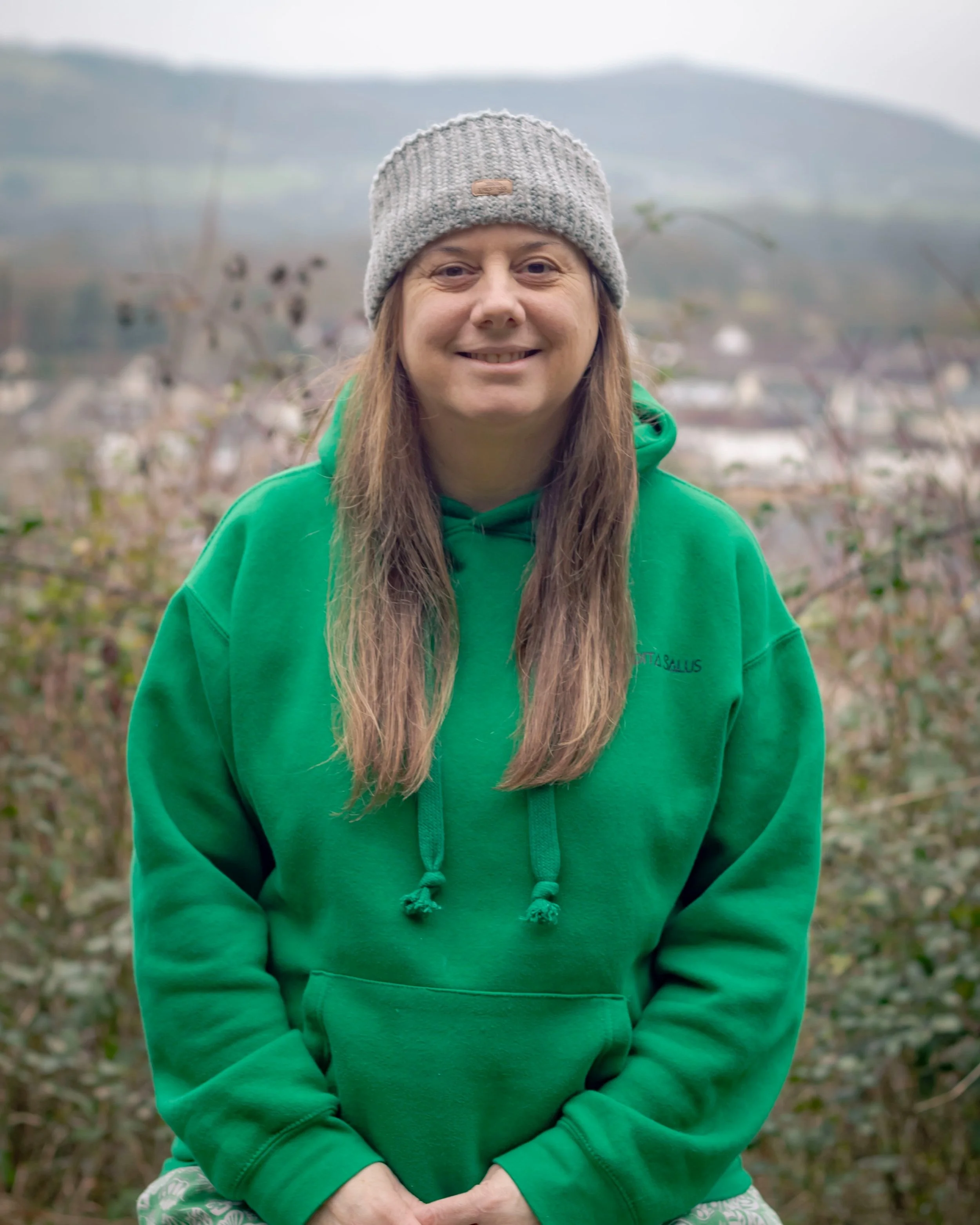 Kim Stone - Mindfulness Teacher:  wearing a grey knit beanie and a green hoodie, standing outdoors with a blurred landscape of trees and distant hills in the background.