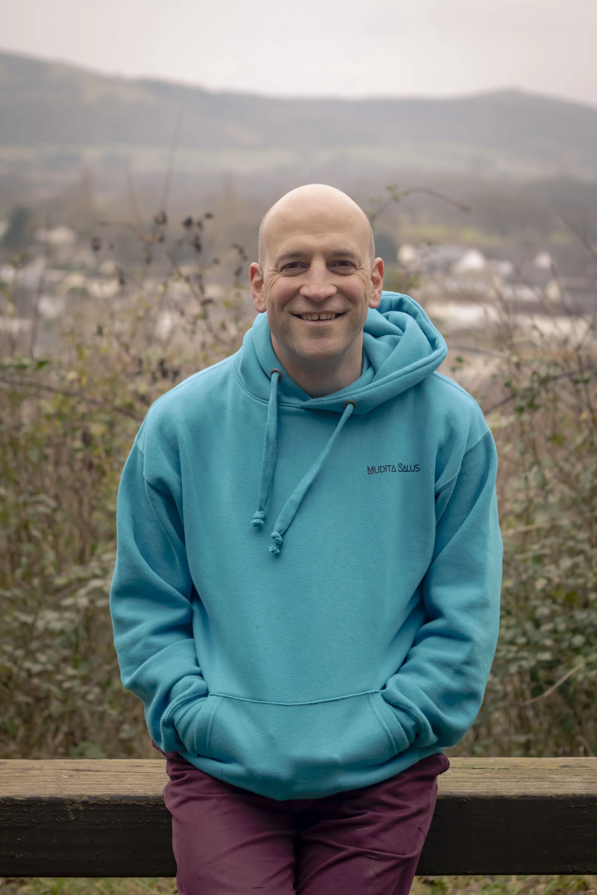 Lee Townsend - Nature & Health Facilitator: in a blue hoodie with hands in pockets, standing outdoors behind a wooden railing, with a blurred hilly landscape and cloudy sky in the background.