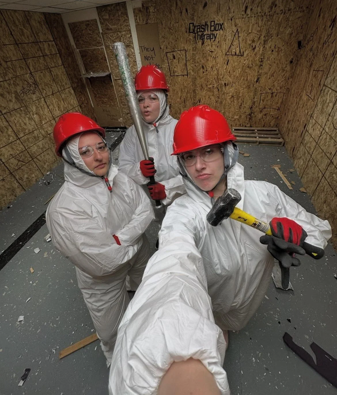 Three women in protective gear with red hard hats and gloves inside a room with unfinished wood walls, holding tools, with debris on the floor, at Crash Box Therapy. 