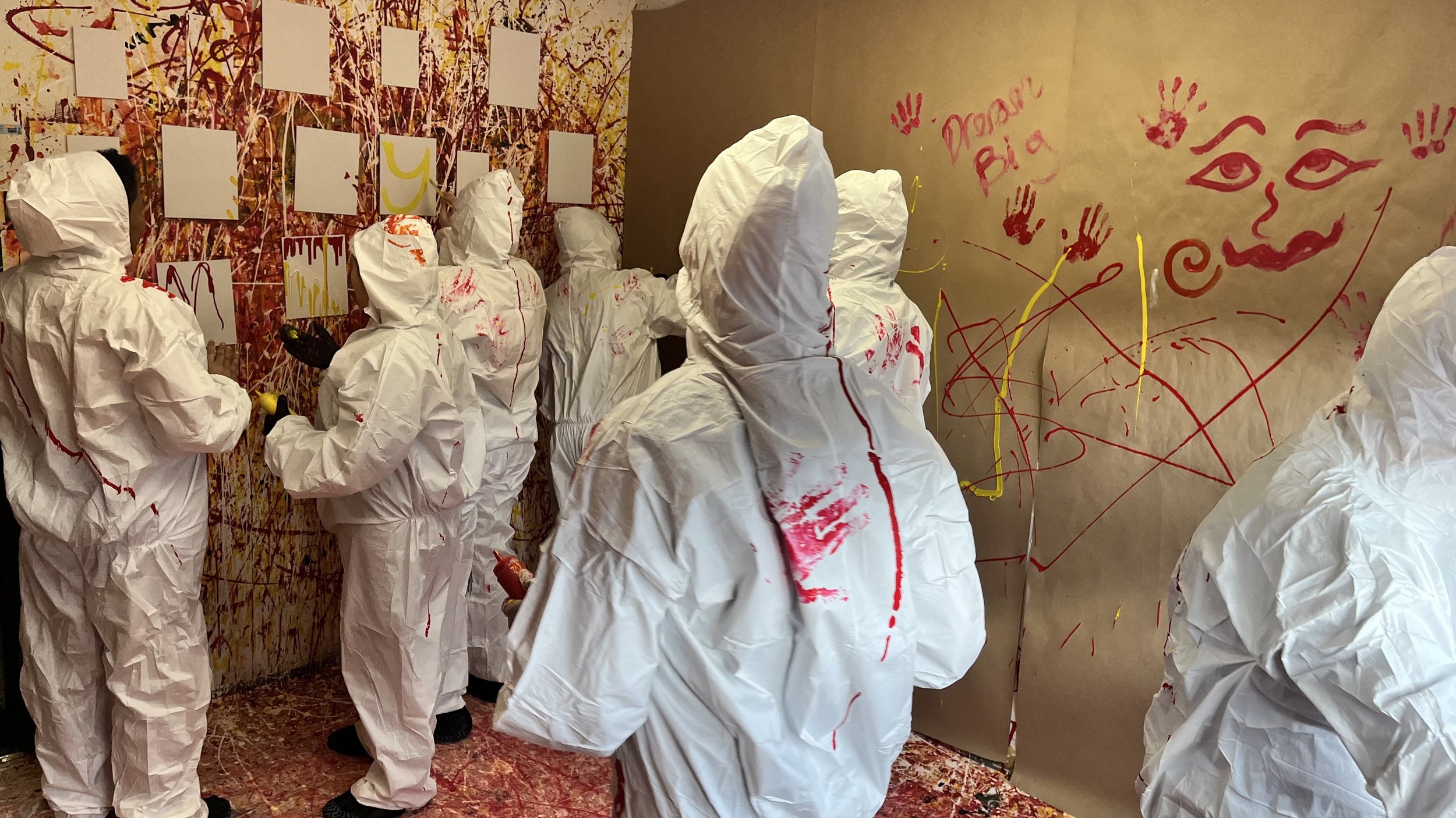 People dressed in white protective suits with red paint splatters are painting and drawing on a wall in an art room.