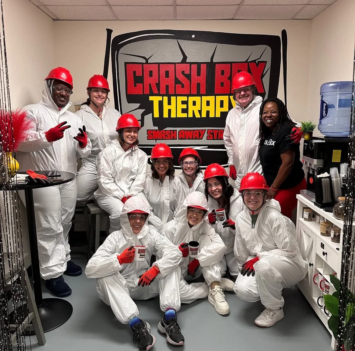Group of people in white coveralls and red safety helmets in a room with a "Crash Box Therapy" logo on the wall, posing for a photo. Corporate Smash 