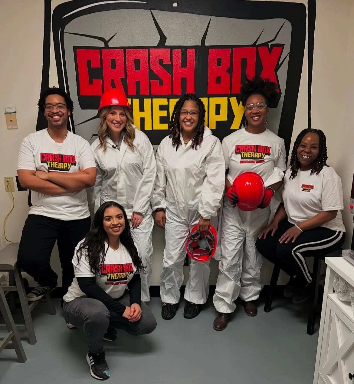 Group of six people, some wearing white coveralls and red hard hats, standing in front of a "Crash Box Therapy" logo.