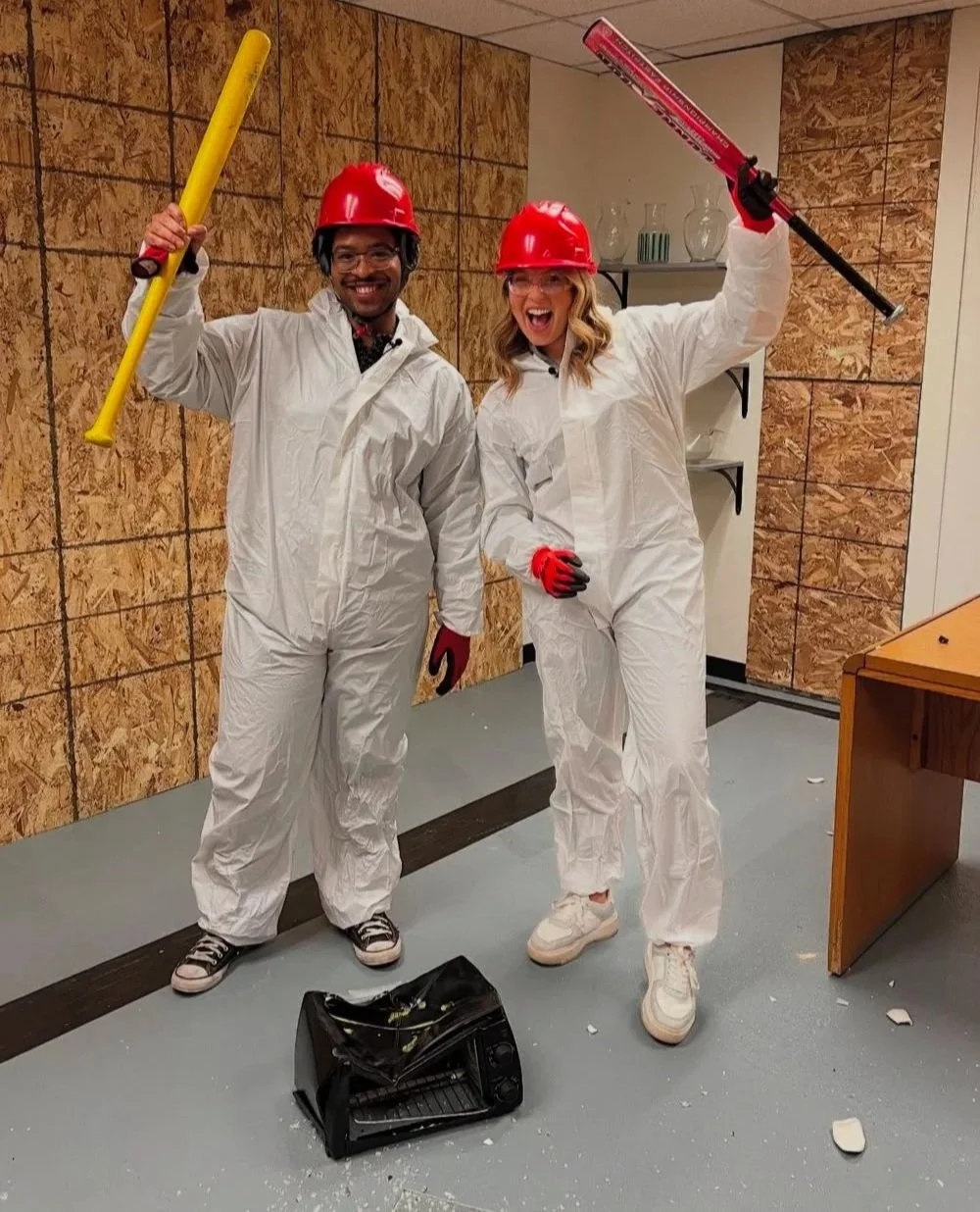 Two people posing in a room wearing white protective suits, red hard hats, and gloves. They are holding colorful baseball bats and smiling. A smashed object is on the floor, suggesting they are in a rage room.